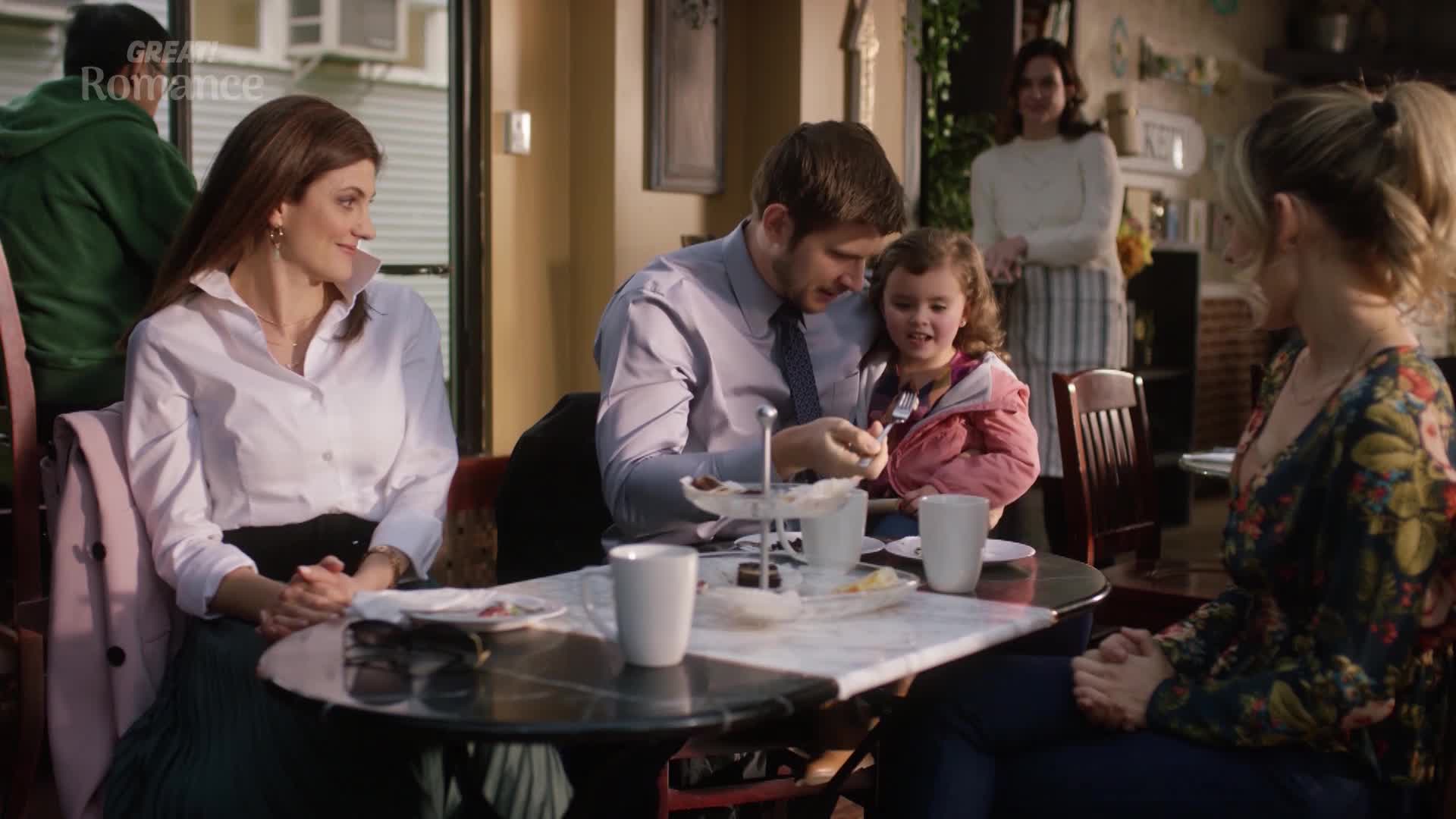 A young man holds a little girl on his lap, both looking at something on a small plate. Across the table, a woman in a white blouse smiles warmly.