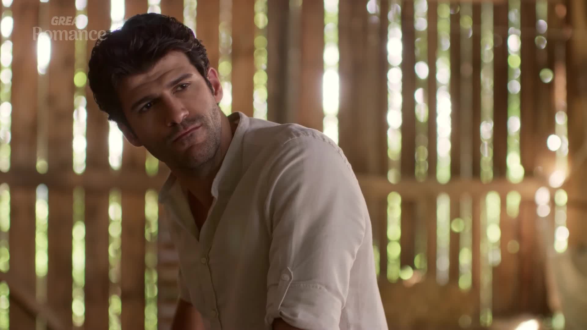 Sunlight streams through the gaps in the wooden barn walls, catching the man's thoughtful gaze. He turns his head, a hint of a smile playing on his lips as he looks off to the side.