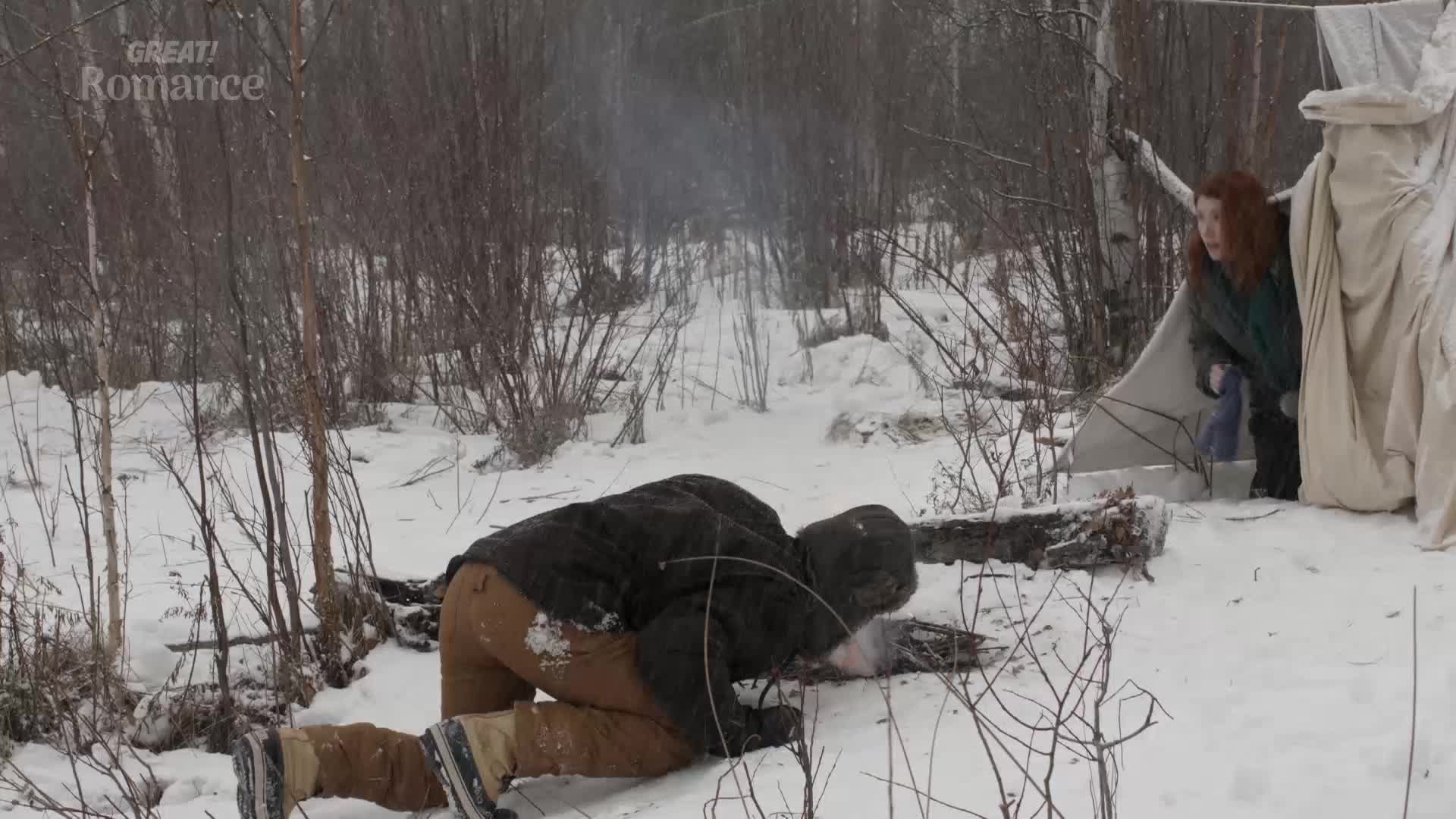 A man in a dark jacket and brown trousers is on his hands and knees in the snow, reaching for something. A woman with red hair peeks out from behind a makeshift shelter, looking alarmed.