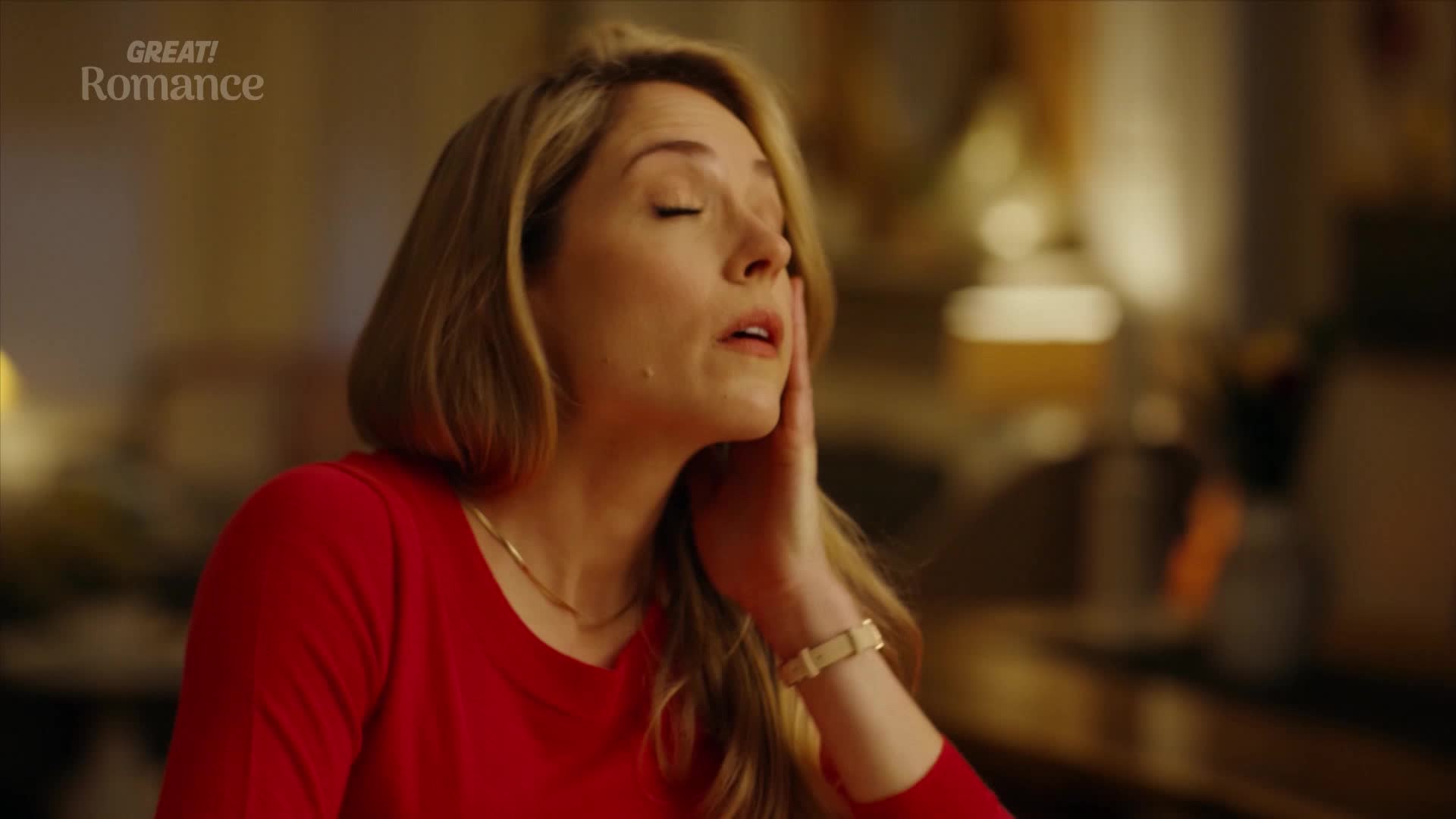 A woman in a red top closes her eyes, her hand gently touching her cheek. The warm, ambient light suggests a cozy evening in, perhaps after a long day.