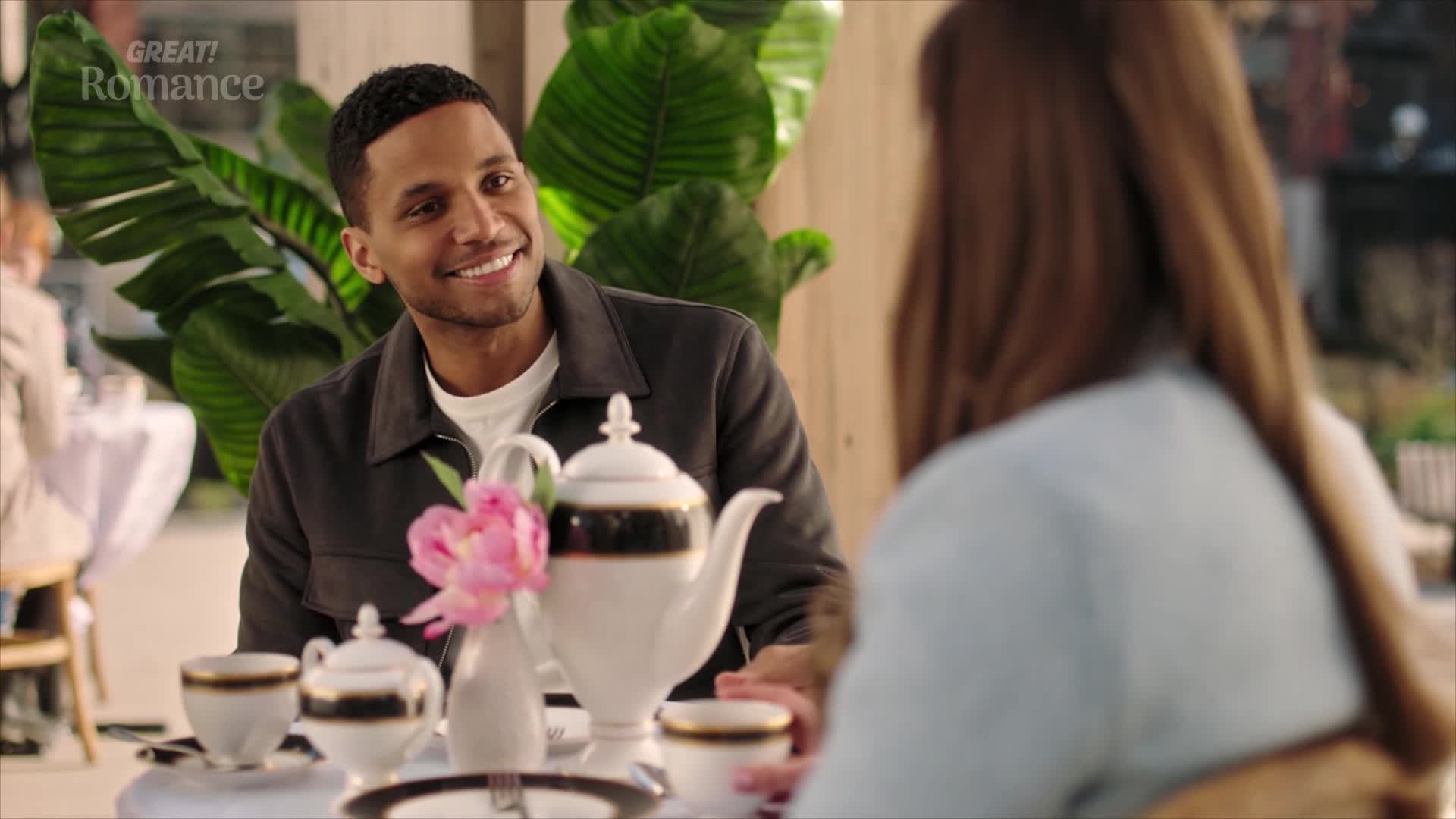 A man with a wide smile looks across a table set for tea. A woman's back is turned, her hair a rich brown, as they share a moment in a sunlit cafe.