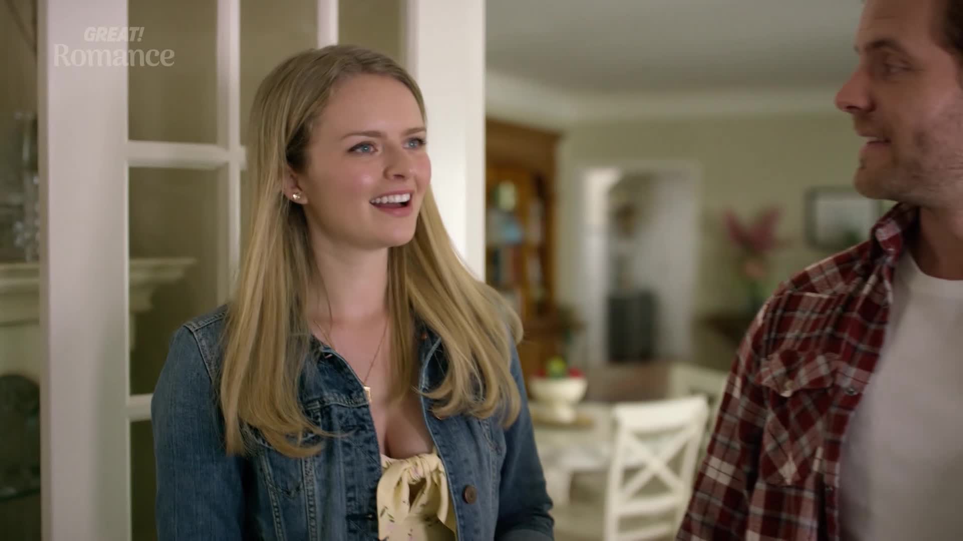A young woman with a bright smile turns towards a man in a red plaid shirt. They stand near a doorway, bathed in soft interior light.