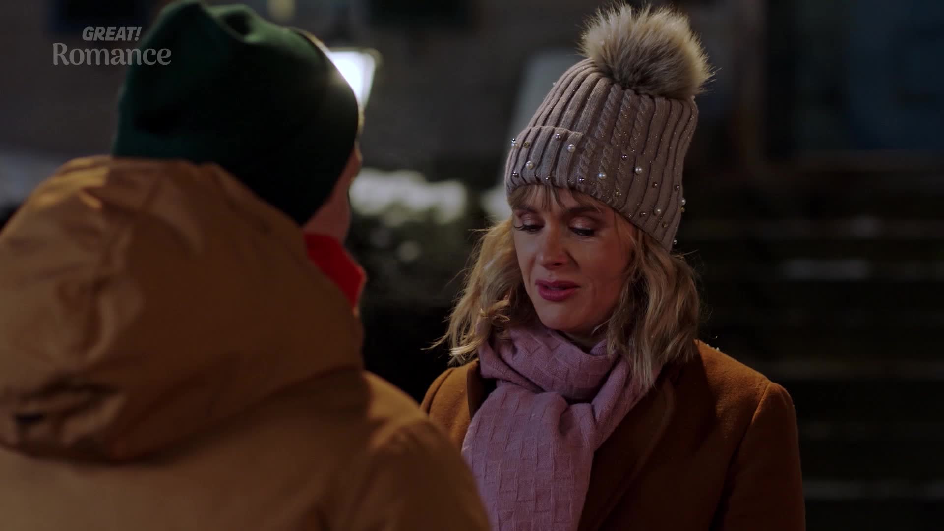A woman in a pale pink scarf and a knitted hat with a pom-pom is looking down, her eyes glistening. Beside her, a man in a tan jacket and a green beanie turns away.