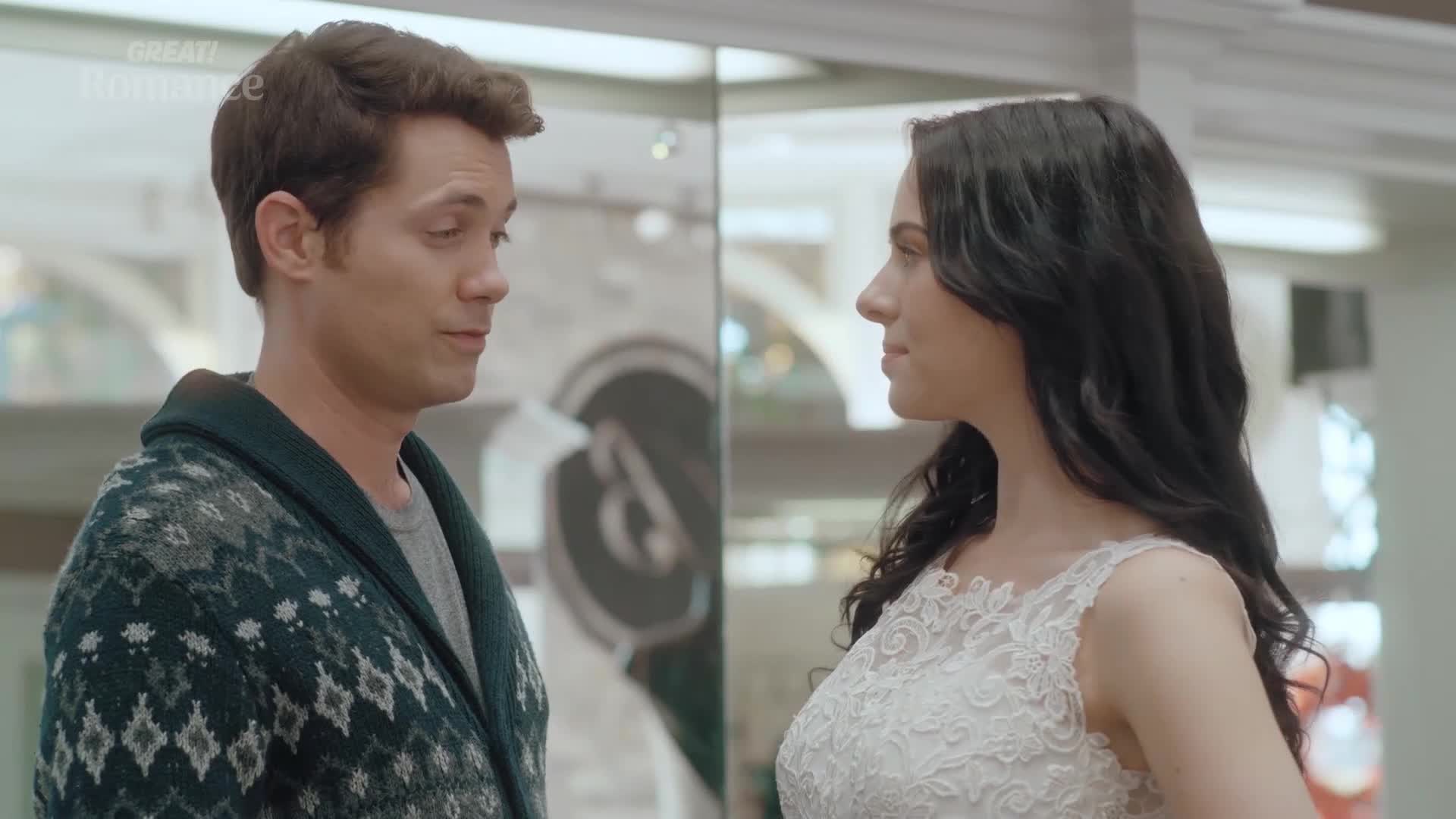 A man in a patterned cardigan looks at a woman in a white lace dress. She turns her head slightly, her dark hair framing her face.