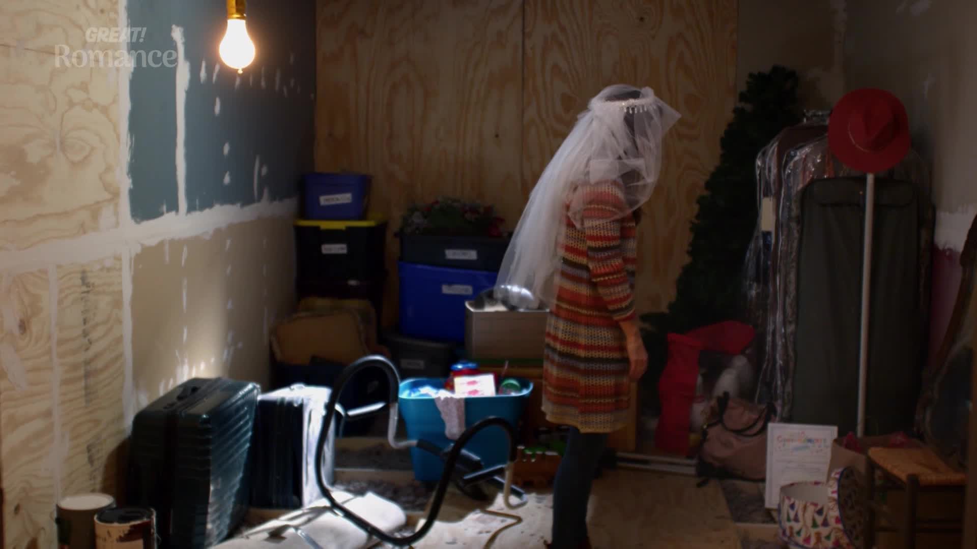 A young woman in a colourful dress and veil stands in a cluttered room. Boxes and luggage are piled high around her, and a bare bulb hangs overhead.