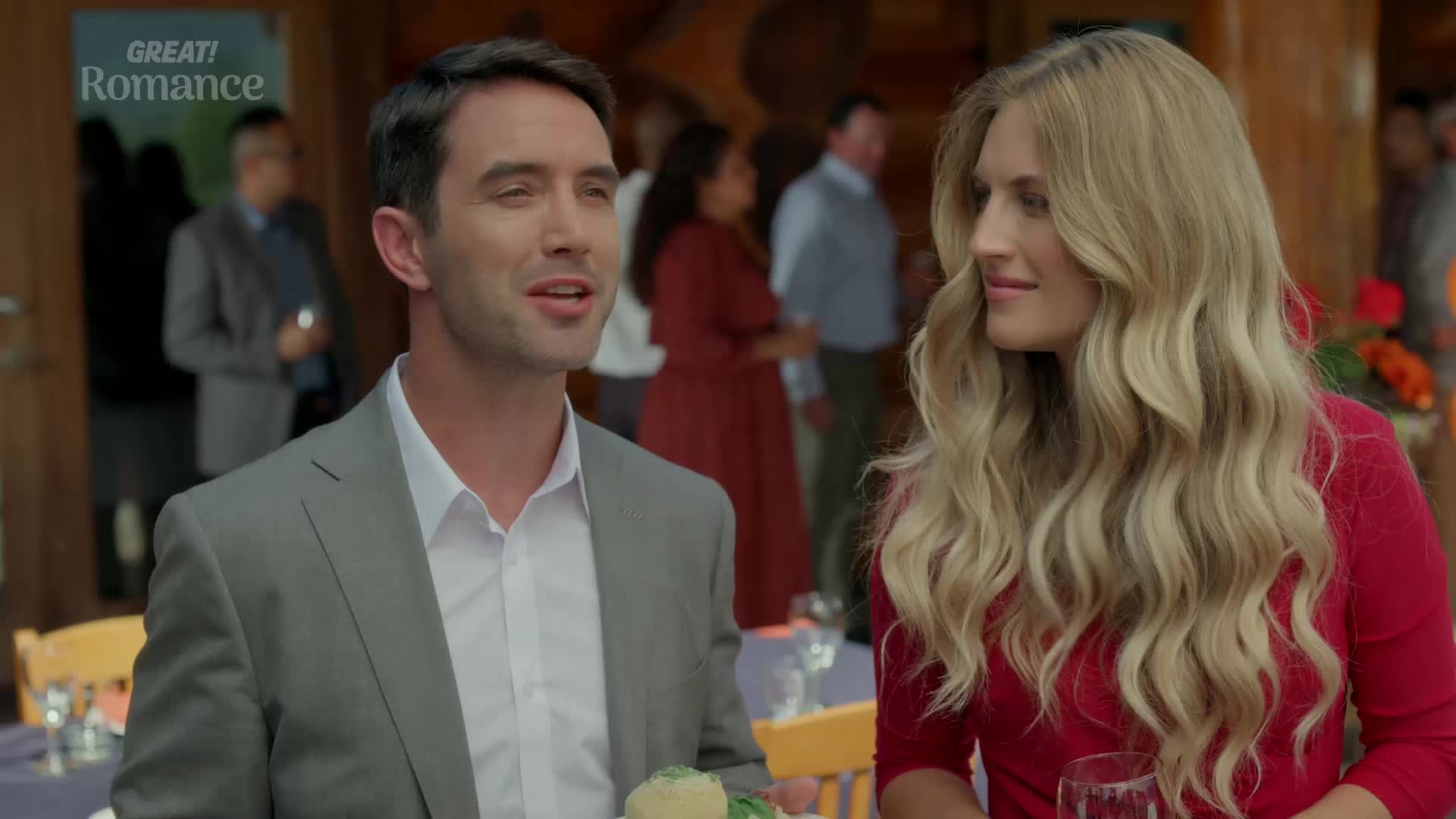 A man in a grey suit leans in, talking to a woman with long blonde hair. They're at some sort of gathering, with other people milling about in the background.