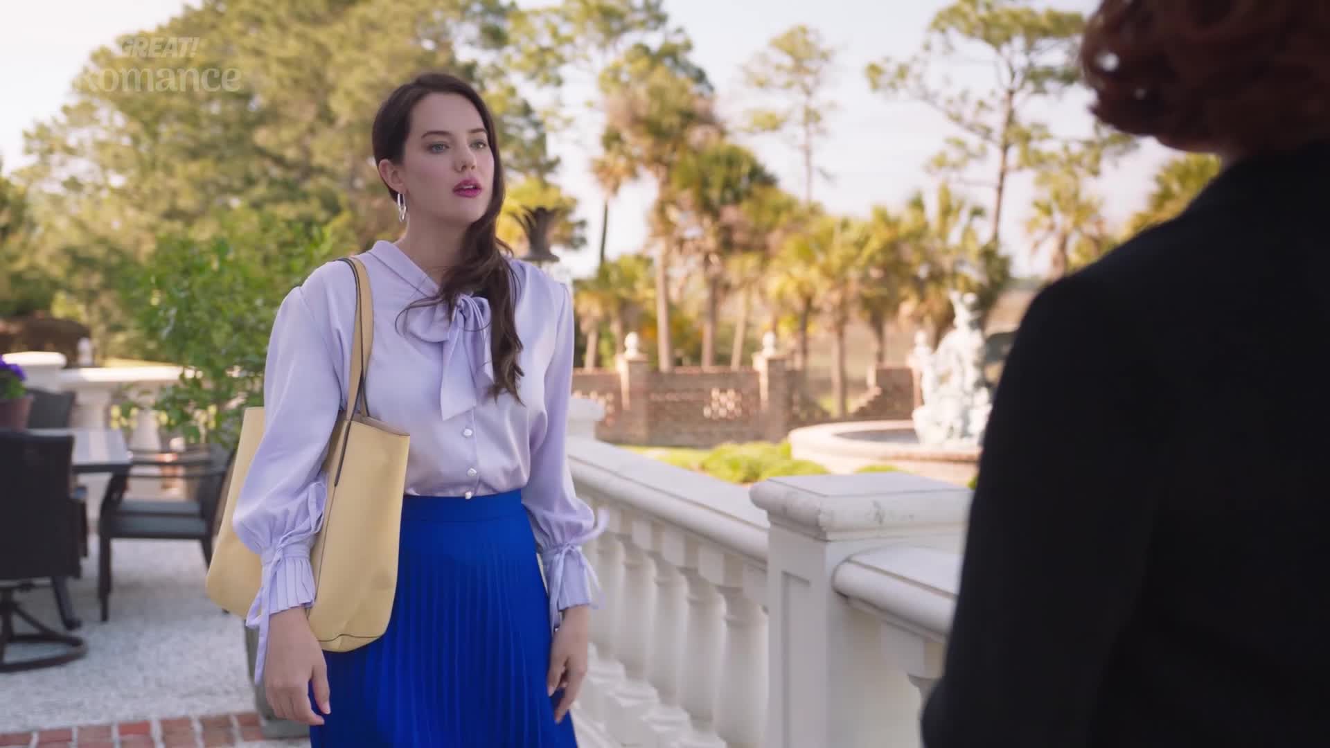 A woman in a lilac blouse and blue skirt stands by a white railing, her expression thoughtful. Behind her, palm trees and an ornate fountain suggest a warm, perhaps Mediterranean, setting.