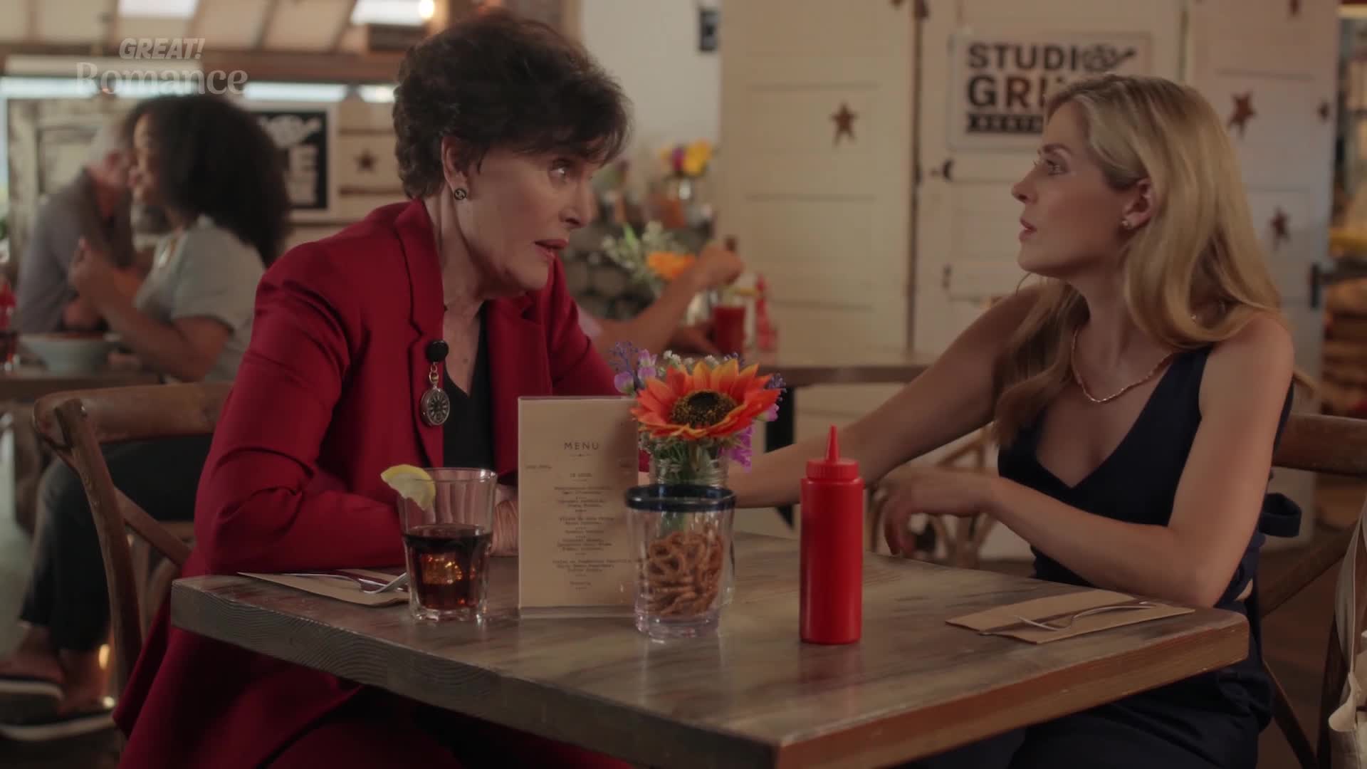 Two women sit at a table in a casual diner, engrossed in conversation. A bright red ketchup bottle stands between them, next to a small vase holding a sunflower and some pretzels.