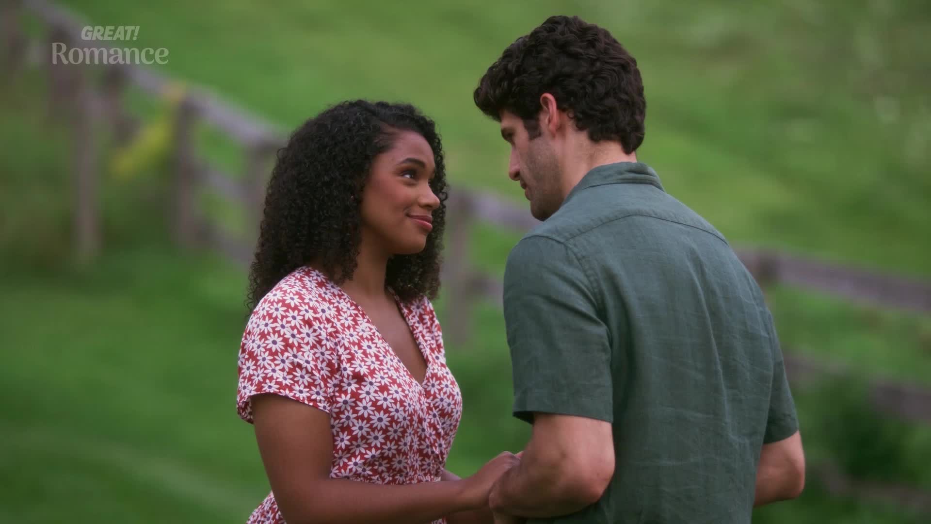 A young woman in a red and white floral dress gazes at a man in a dark green shirt. They stand close, their hands clasped, amidst rolling green fields typical of the British countryside.