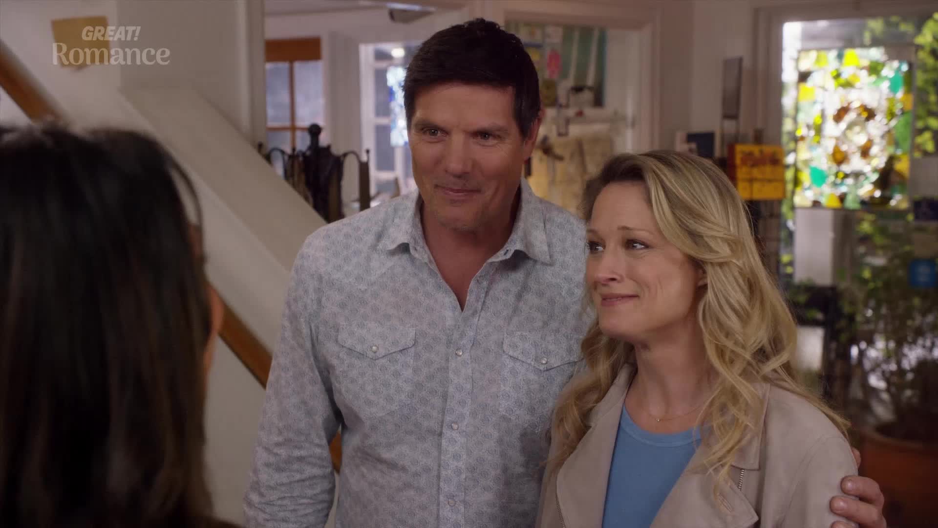 A man in a patterned shirt and a woman with blonde hair stand close, their gazes directed off-frame. The woman smiles softly, a hint of warmth in her expression.