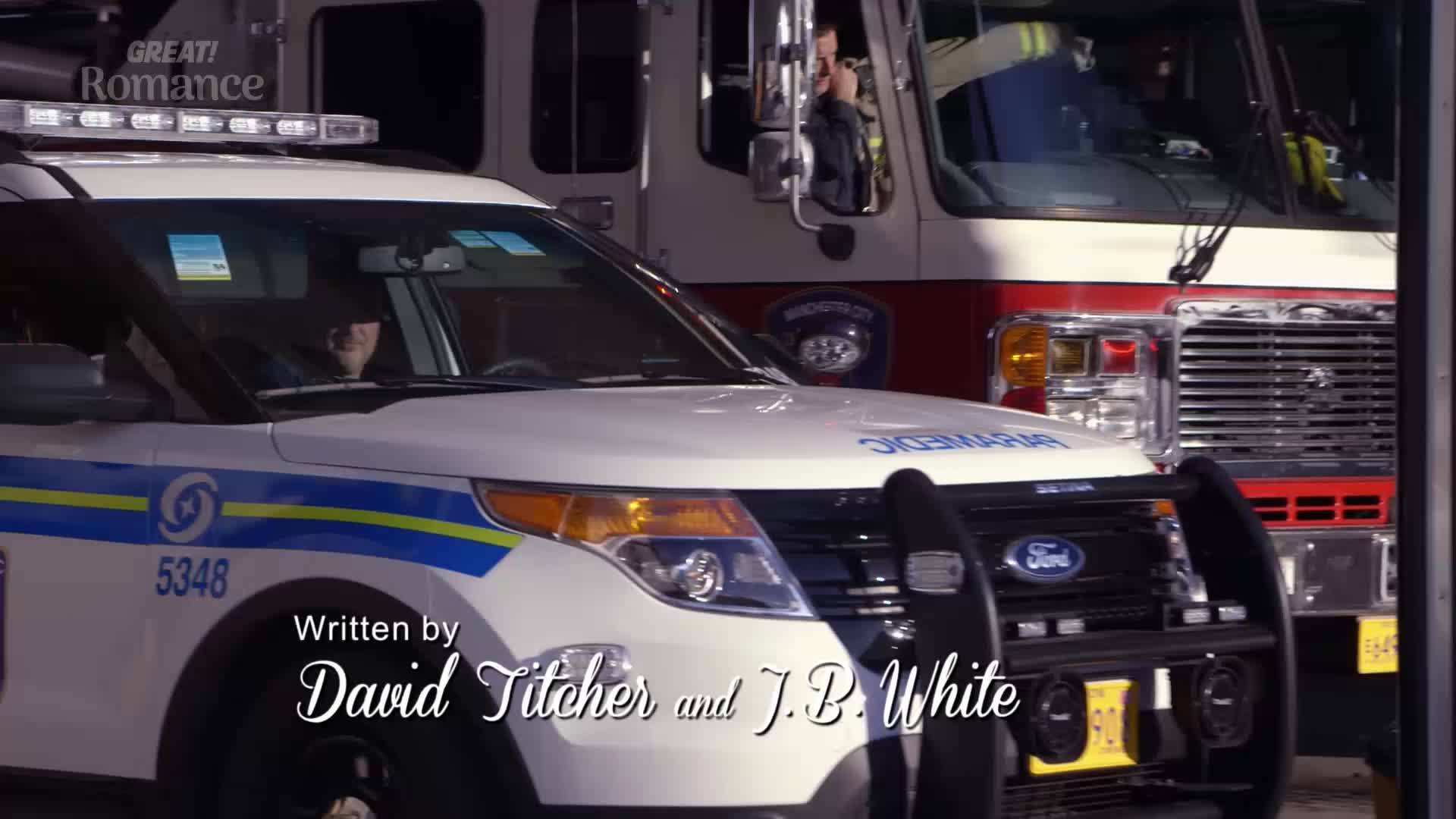 A white police car, lights flashing, sits in front of a red fire truck. A person in uniform is visible behind the steering wheel of the police car.