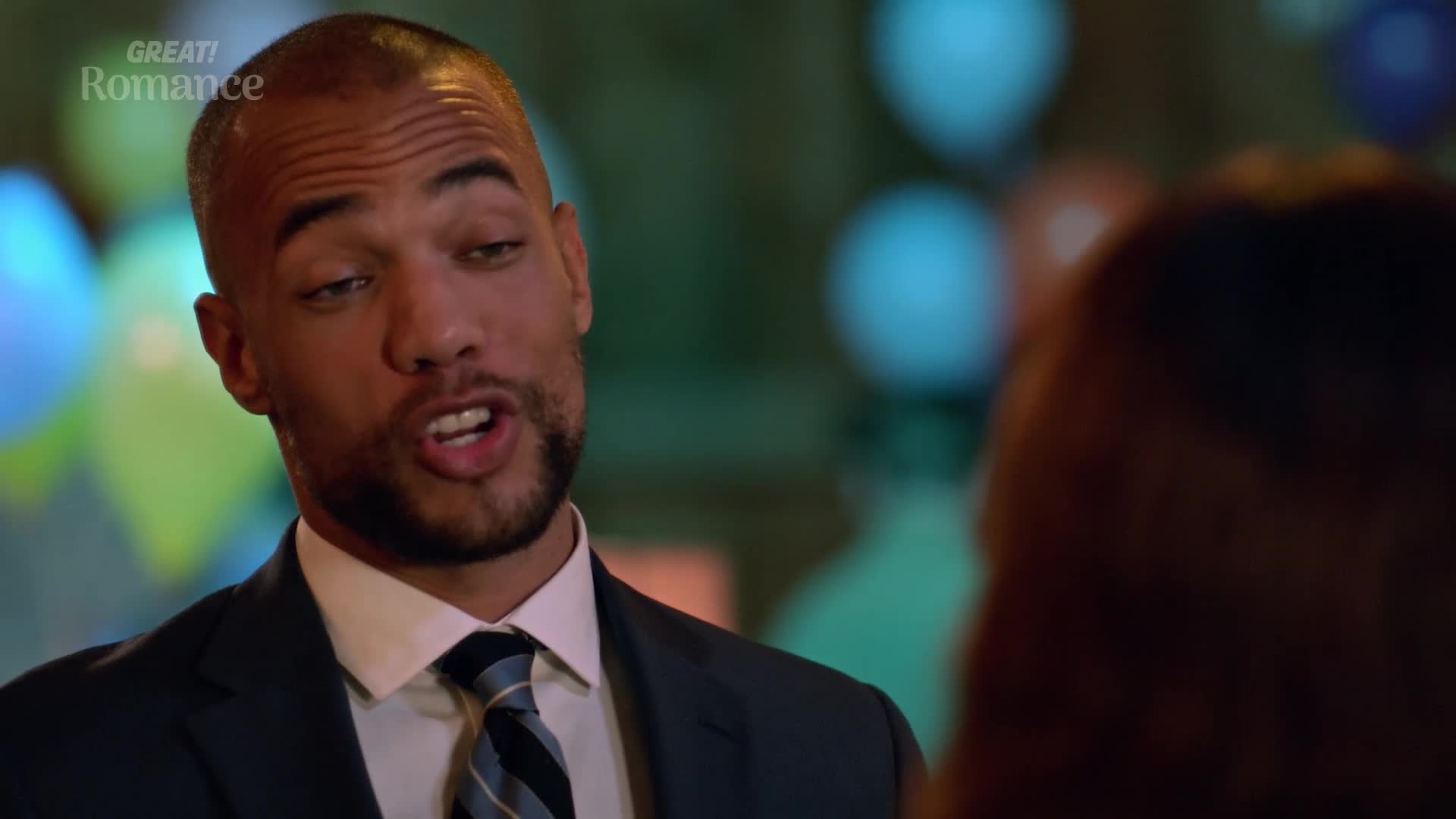 He's leaning in, talking intently, his tie a sharp contrast to the soft glow of balloons behind him. This feels like a moment from a GREAT! Romance, perhaps during a party in the UK.