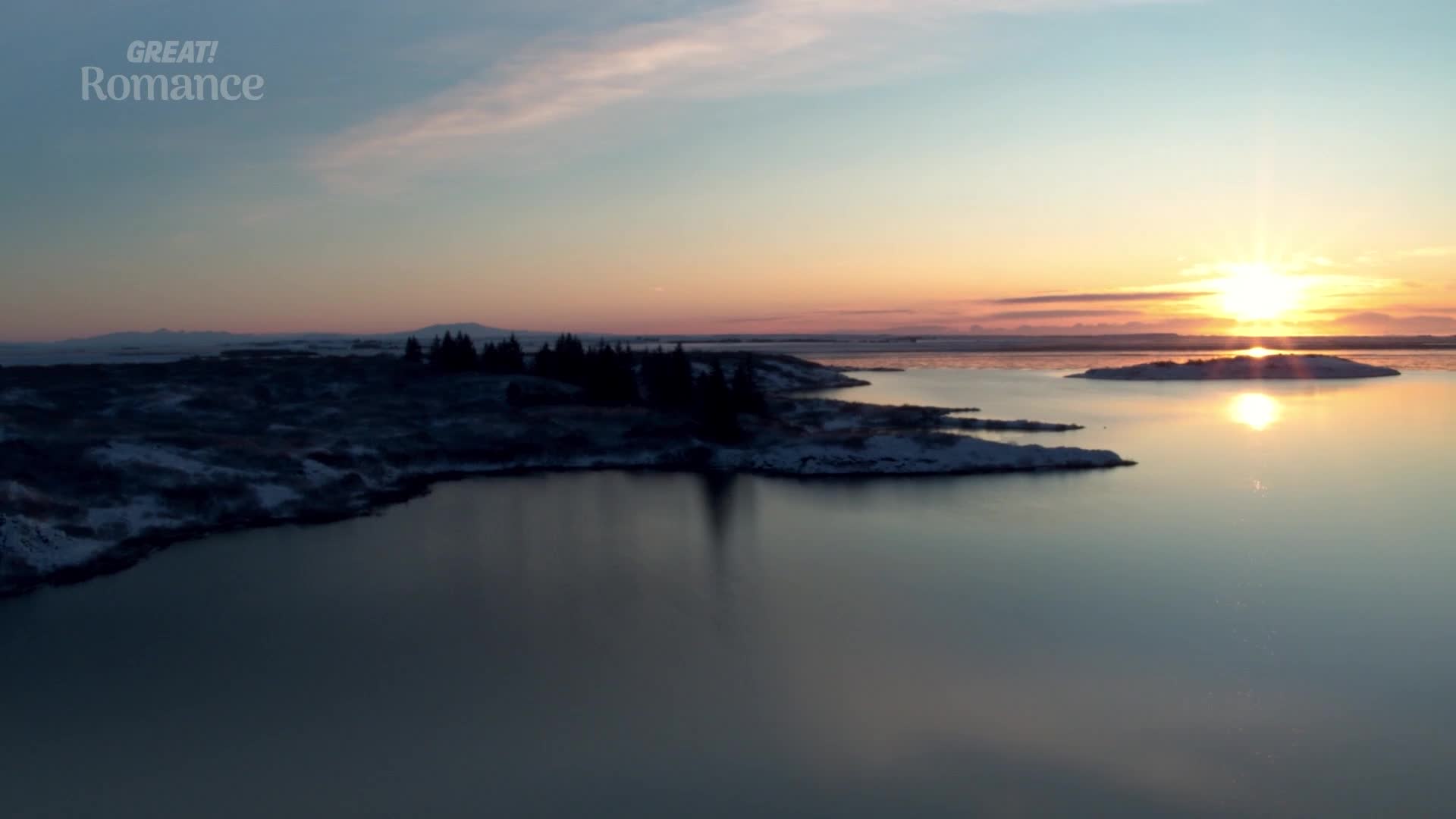 The sun dips low, casting a warm glow over a tranquil lake. Snow-dusted land and dark trees hug the water's edge as the sky blushes pink and orange.