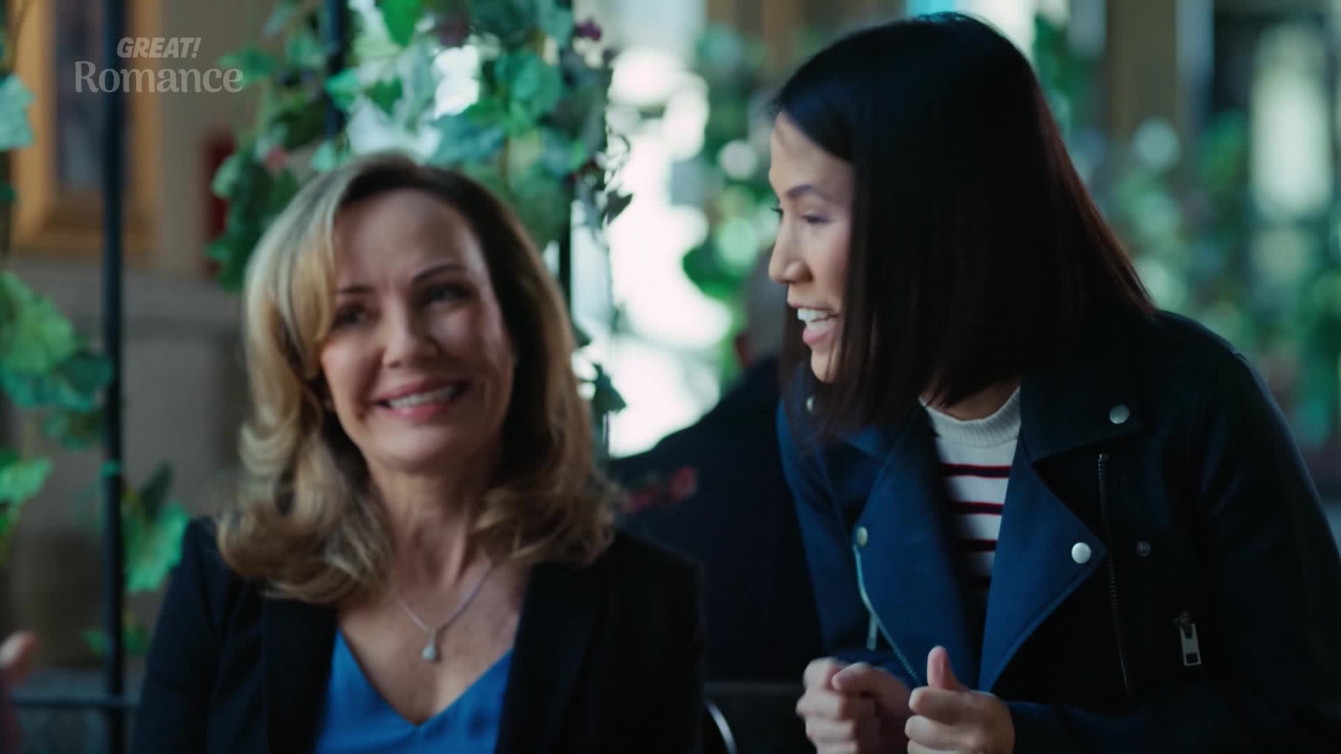 Two women are sharing a moment of laughter over coffee. The one with blonde hair, wearing a blue top and blazer, beams as the other, in a dark jacket, leans in to speak.