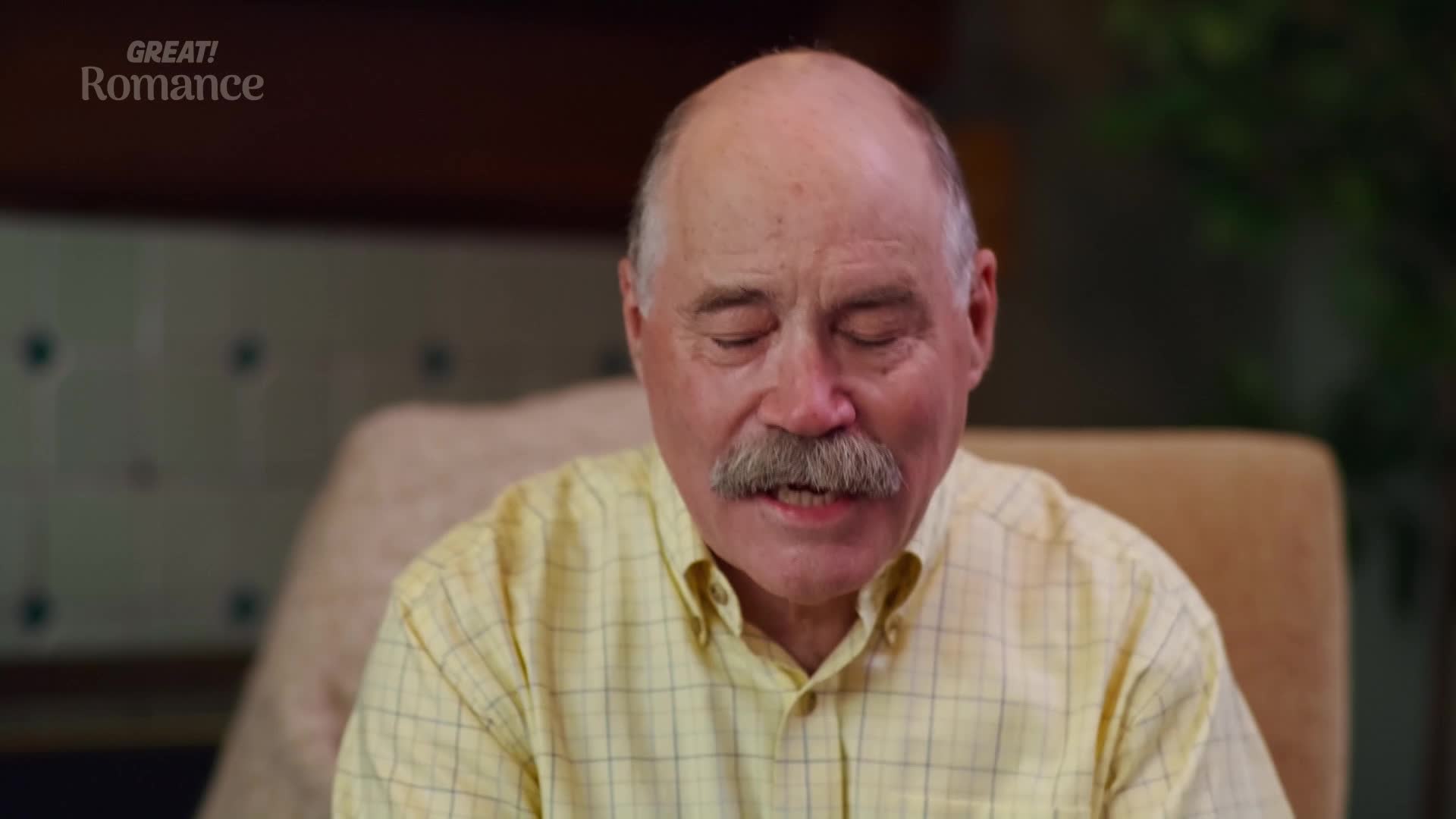 A man with a thick mustache looks down, his eyes closed. He's wearing a pale yellow checkered shirt, and the "GREAT! Romance" logo is visible in the top left corner.