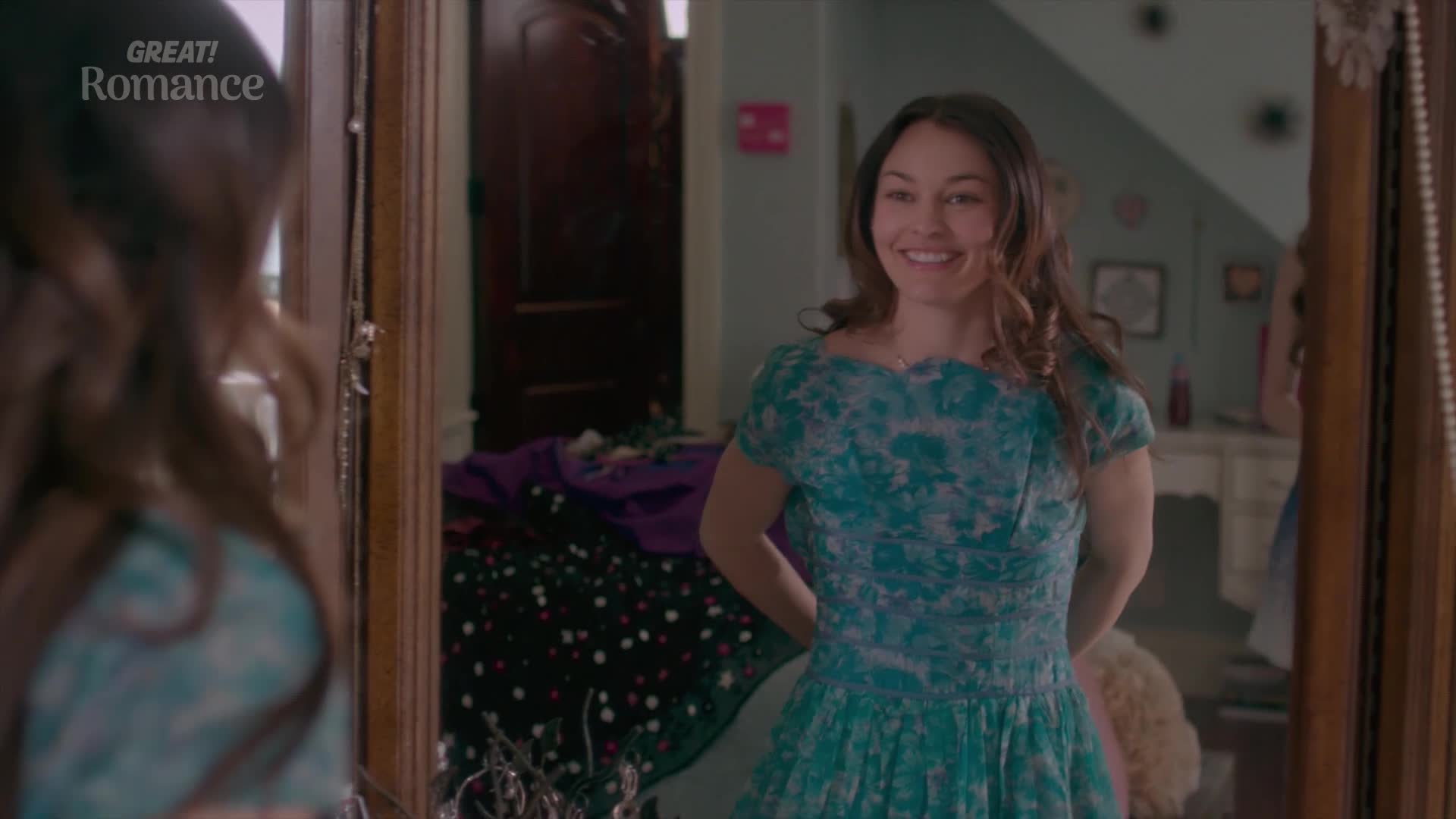 She's admiring herself in the mirror, a wide smile on her face as she twirls in a light blue, patterned dress. The room behind her is a bit of a mess, with clothes piled on the floor.