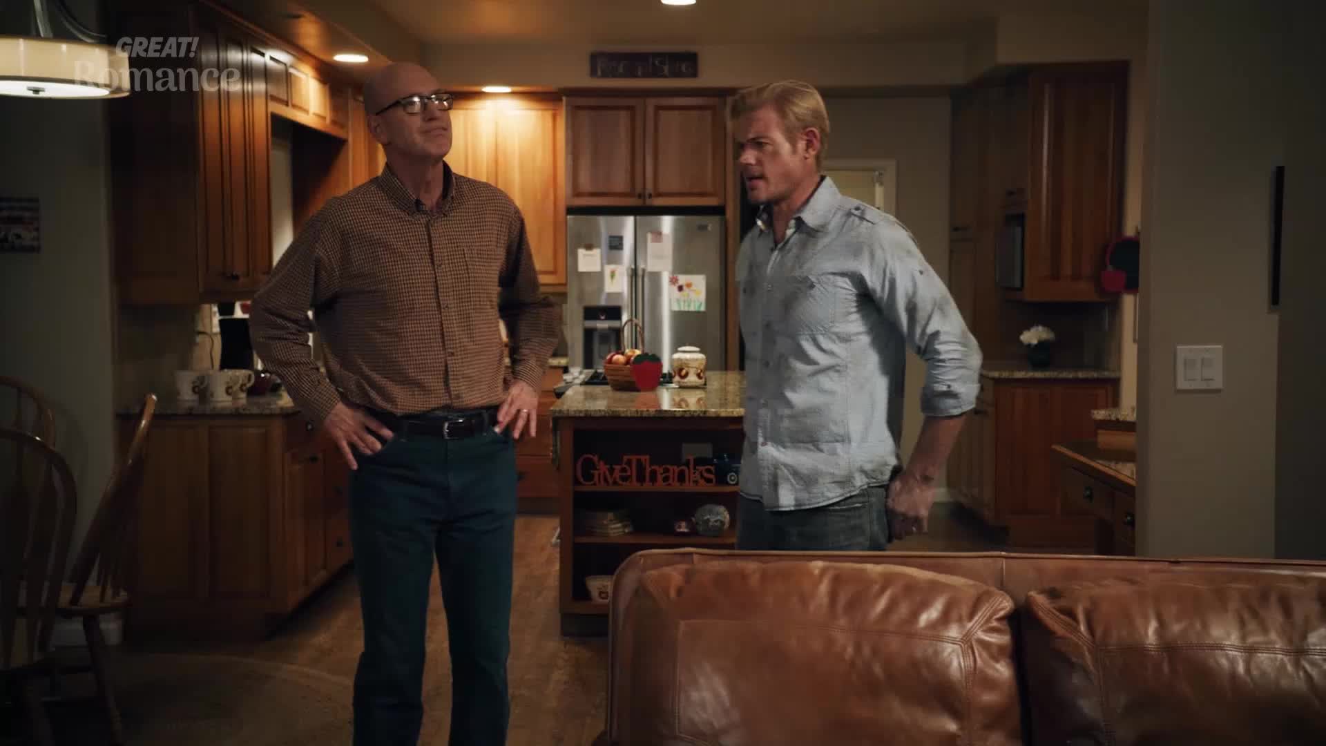 Two men stand in a kitchen, one in a patterned shirt and the other in a blue button-down. A leather sofa sits in the foreground.