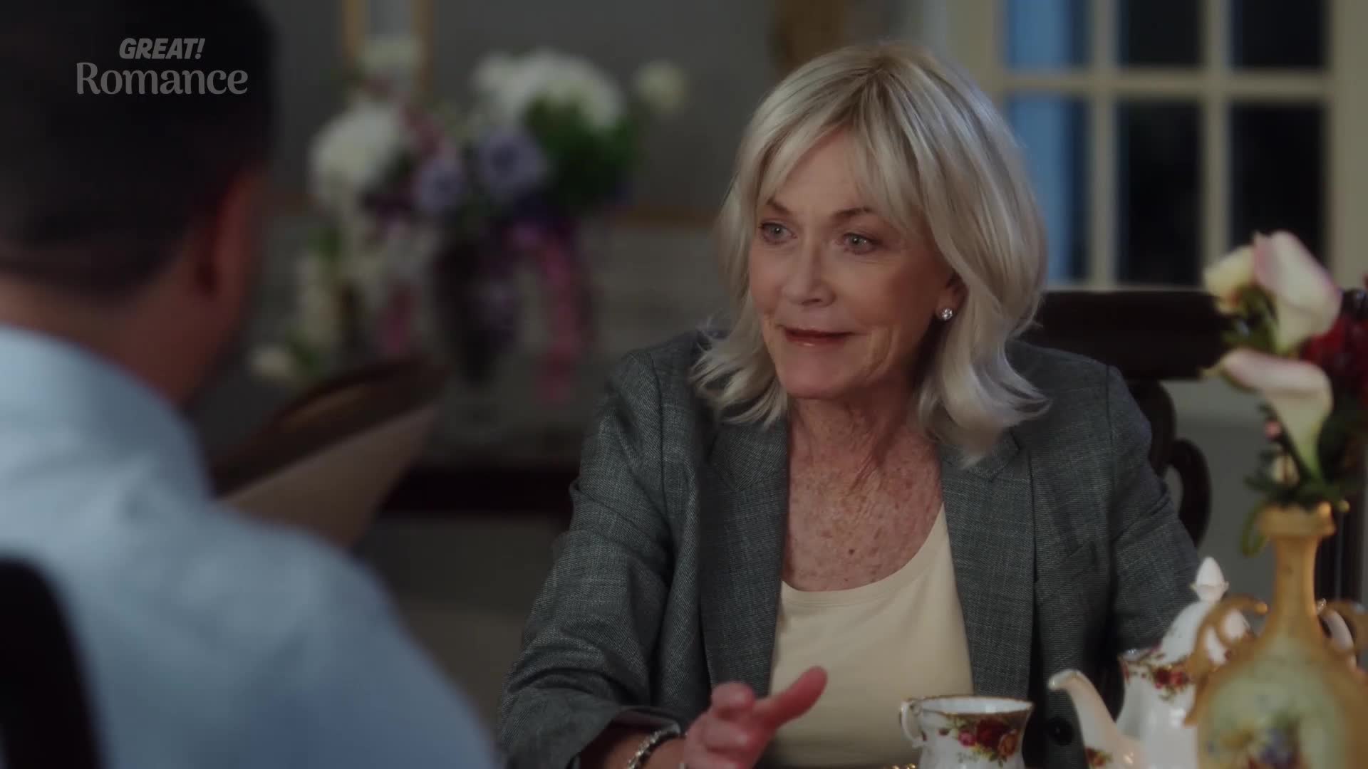 A woman with blonde hair, wearing a grey blazer, gestures as she speaks. She's seated at a table with a delicate teacup, a scene that feels like something from a charming British drama.