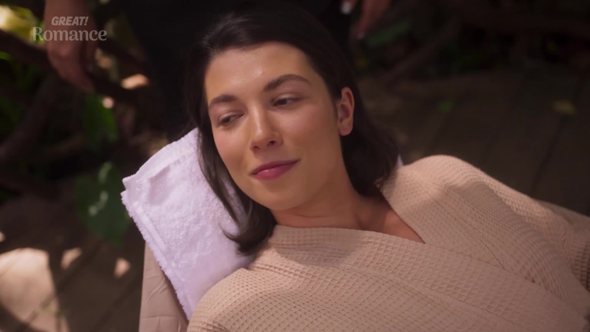 A woman reclines on a towel-covered surface, her gaze soft. She wears a textured robe, hinting at a relaxing spa moment.