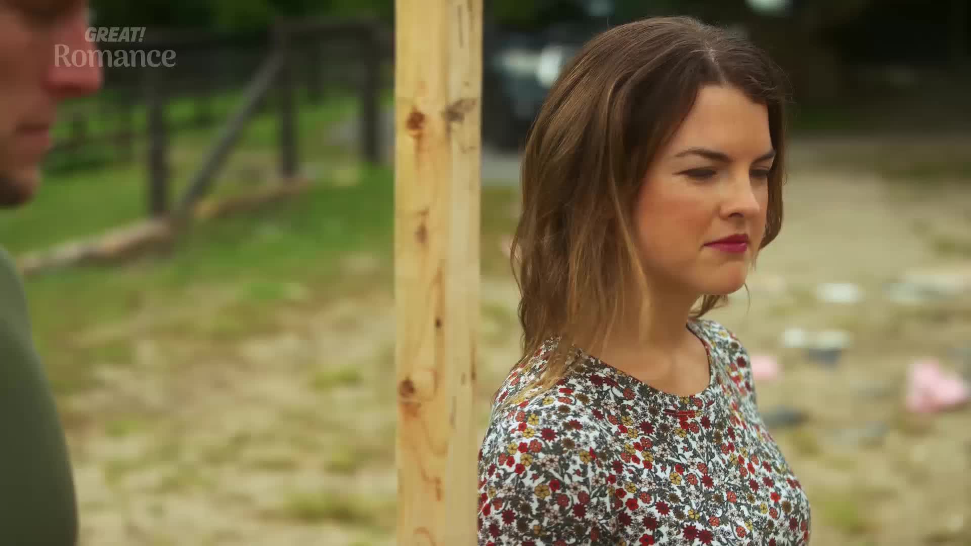A woman in a floral dress stands near a wooden post, her gaze fixed on something out of frame. The background suggests a construction site, perhaps in the English countryside.
