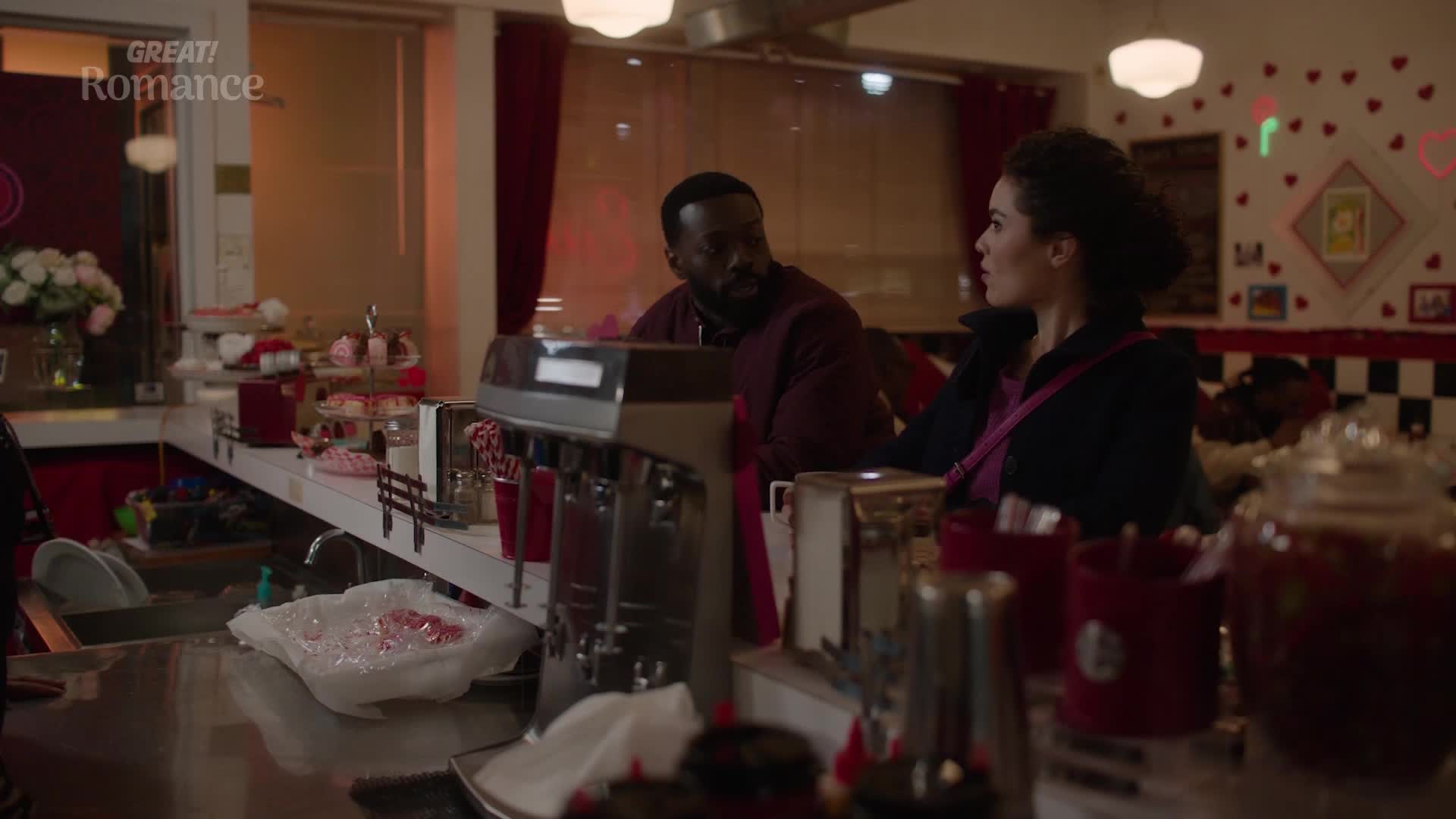 A man and a woman sit at a diner counter, bathed in the warm glow of the interior. The walls are adorned with a playful pattern of red hearts, hinting at a romantic setting.
