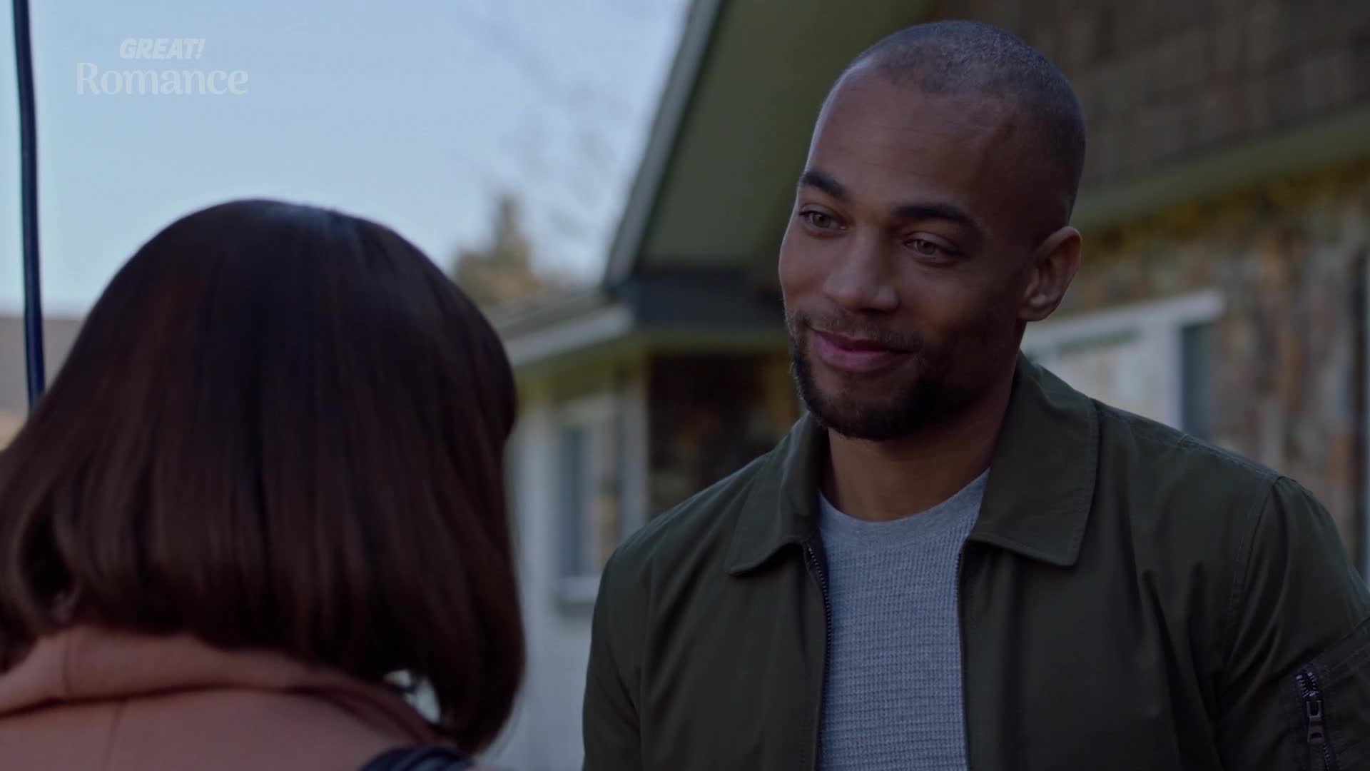 A man with a warm smile looks at a woman whose back is to me. He's wearing a green bomber jacket over a grey t-shirt, and the backdrop suggests a quiet afternoon in the English countryside.