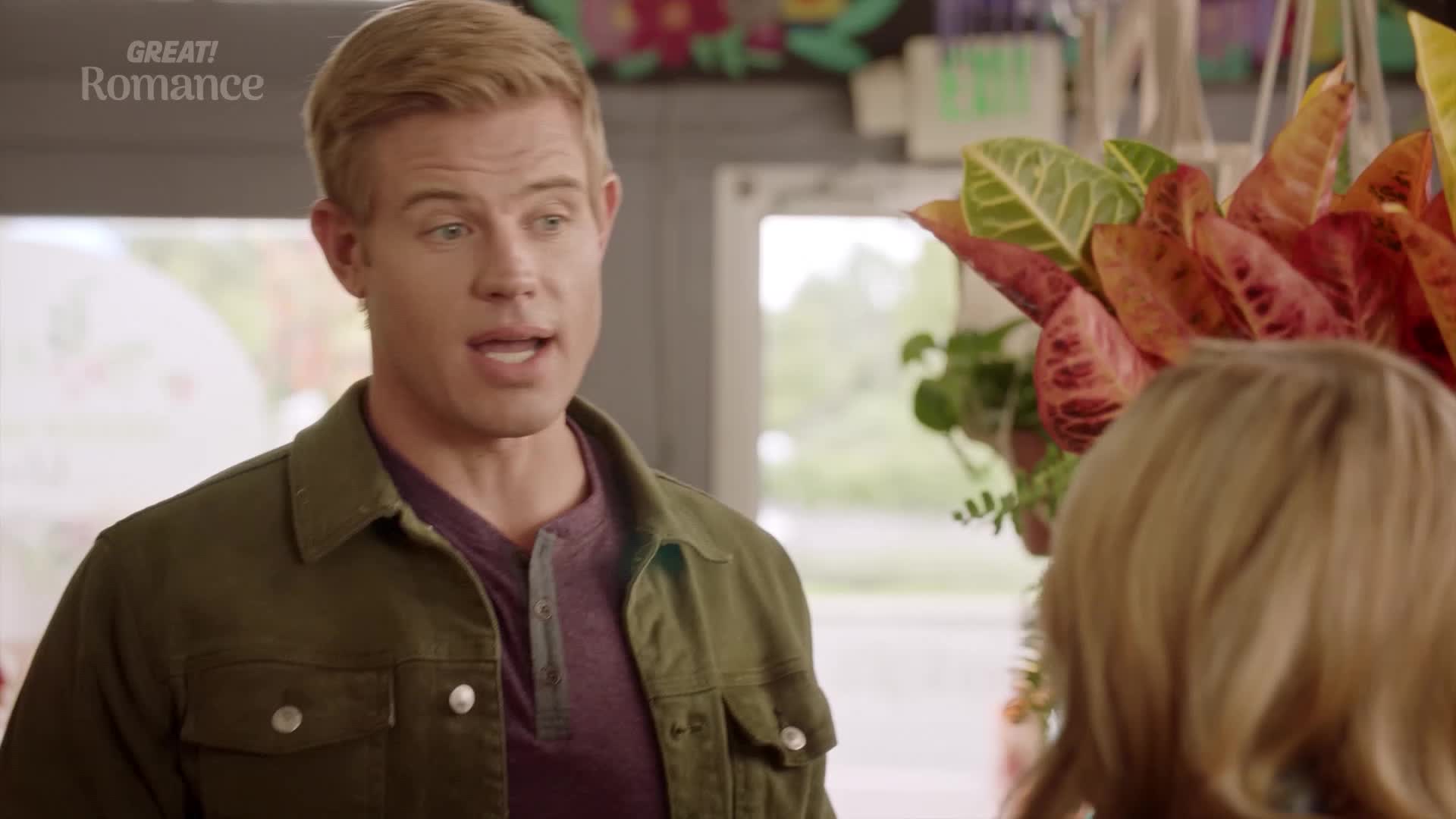 A young man with bright eyes gestures with his hands as he speaks. He's wearing a green jacket over a purple shirt, standing in what looks like a shop filled with plants.