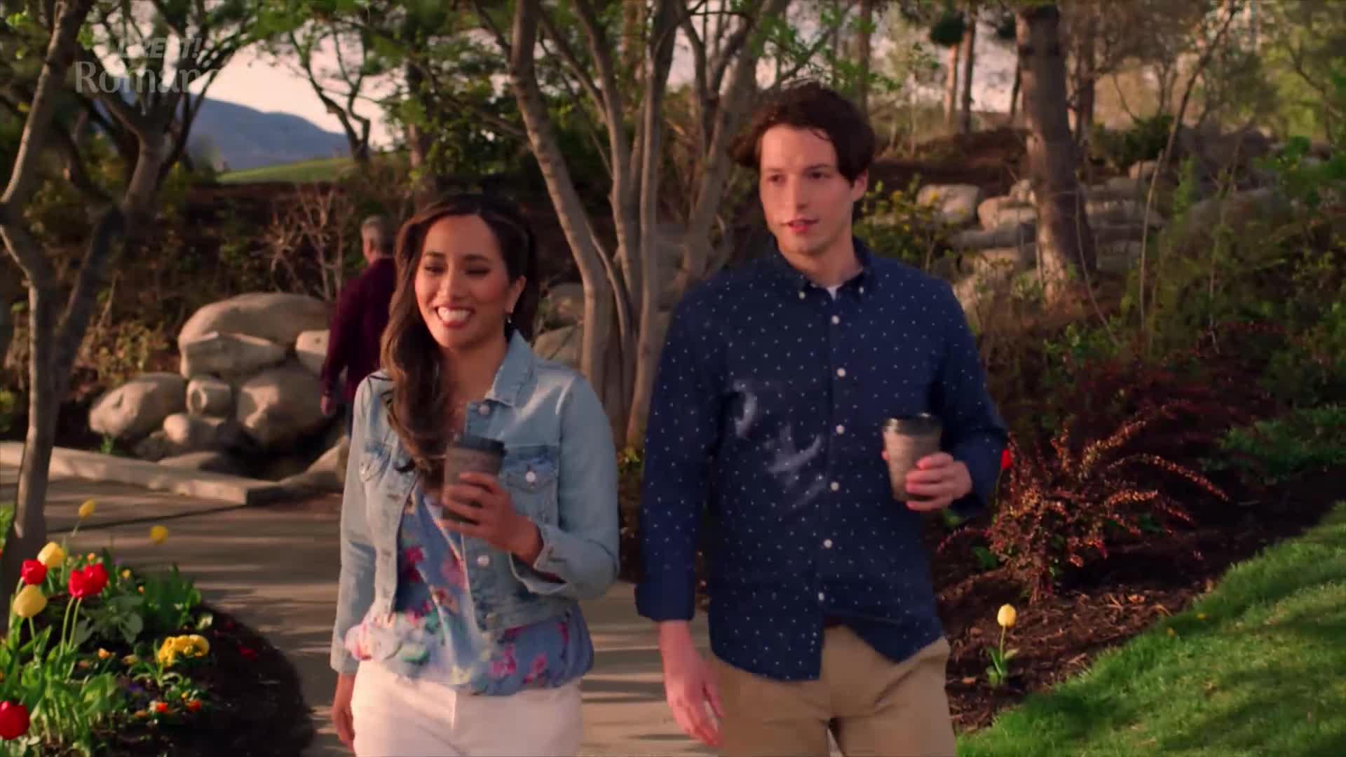 A couple strolls along a garden path, each holding a takeaway coffee cup. The woman, in a denim jacket, smiles warmly as they walk side-by-side.