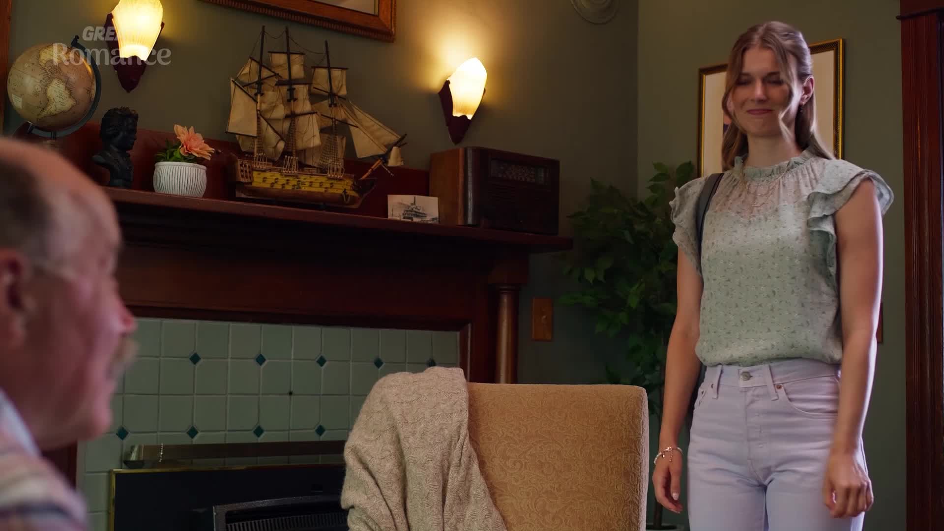 A young woman in a floral blouse and light purple trousers stands smiling, a backpack slung over her shoulder. An older man, his face turned away, sits in the foreground.