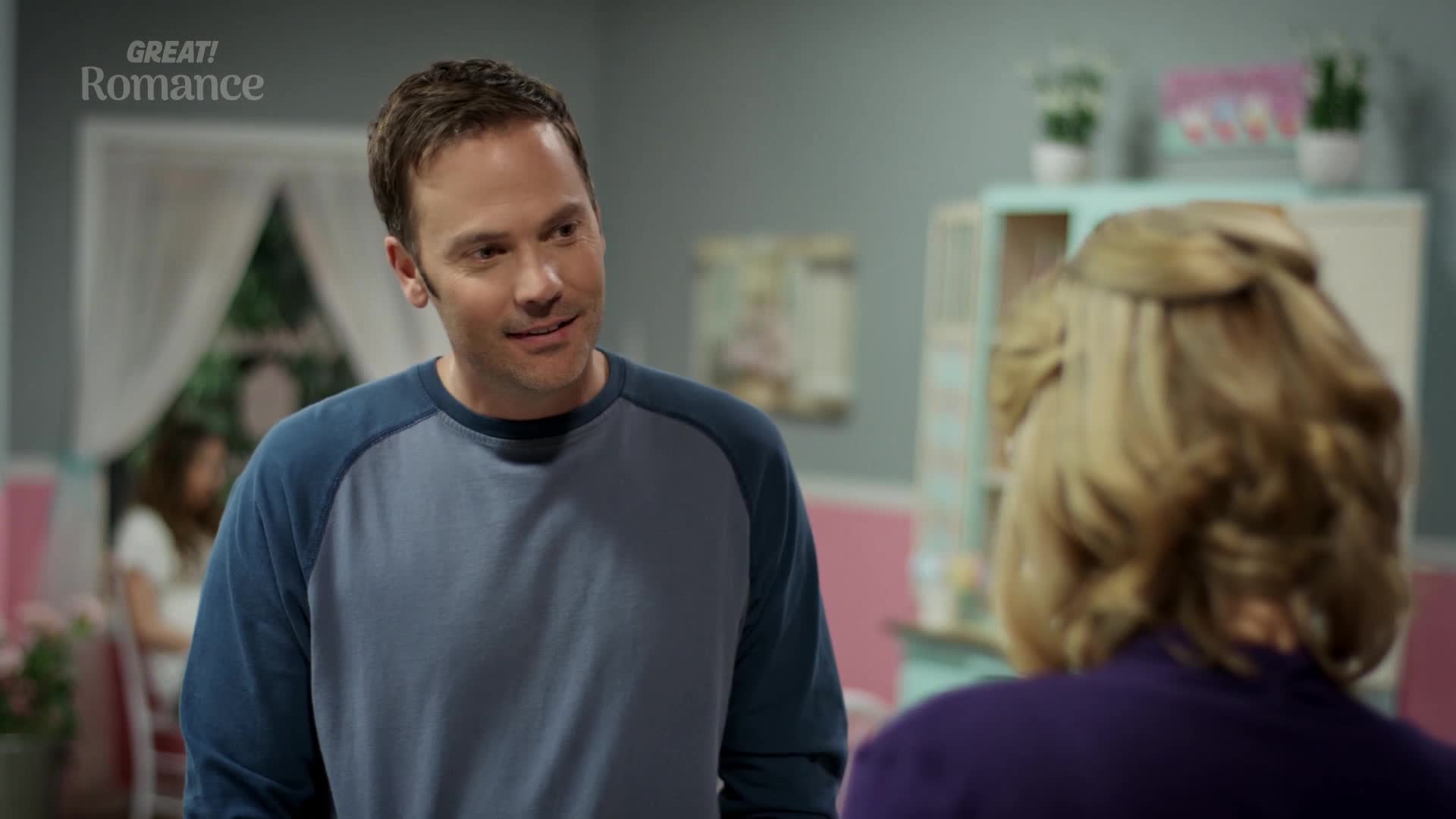 A man with short brown hair, wearing a blue and grey raglan shirt, smiles warmly at a woman whose blonde hair is styled up. Behind them, a young girl sits at a table in a room decorated with pastel colours.