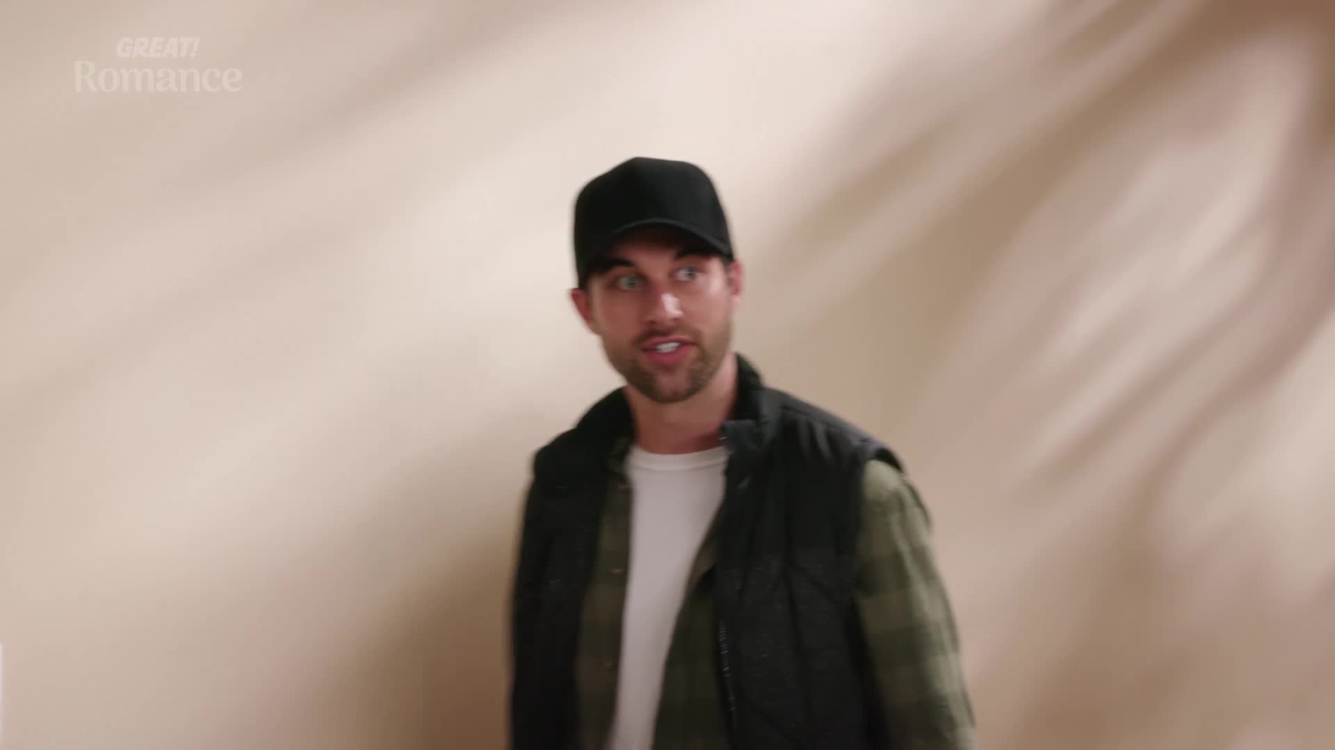 A man in a black baseball cap and a dark vest over a plaid shirt turns his head, speaking. The background is a soft, warm beige with subtle shadows.