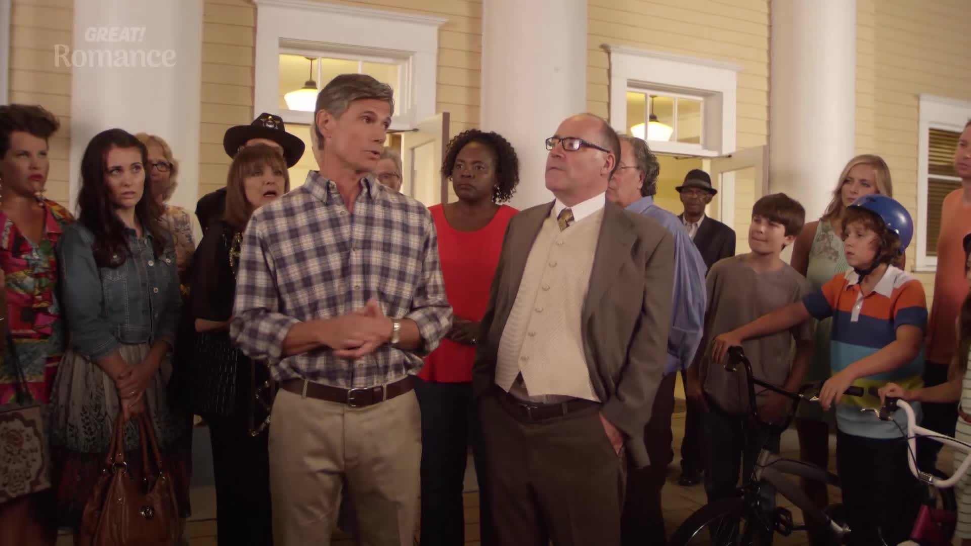 A man in a plaid shirt addresses a crowd gathered outside a building. Two boys stand with their bicycles, watching the proceedings.