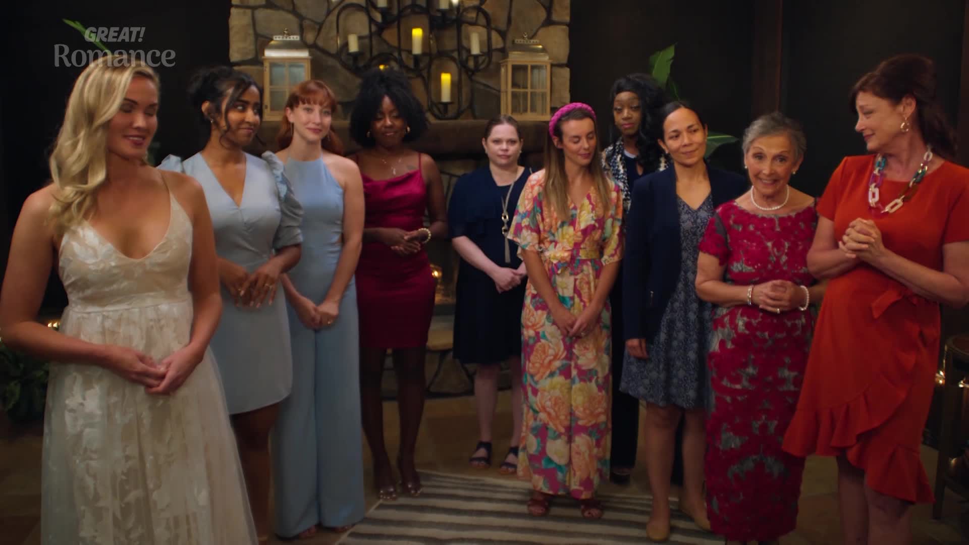 A line of women stands facing forward, dressed for an occasion. The woman on the far left wears a white lace gown, while others are in various colourful dresses and jumpsuits.