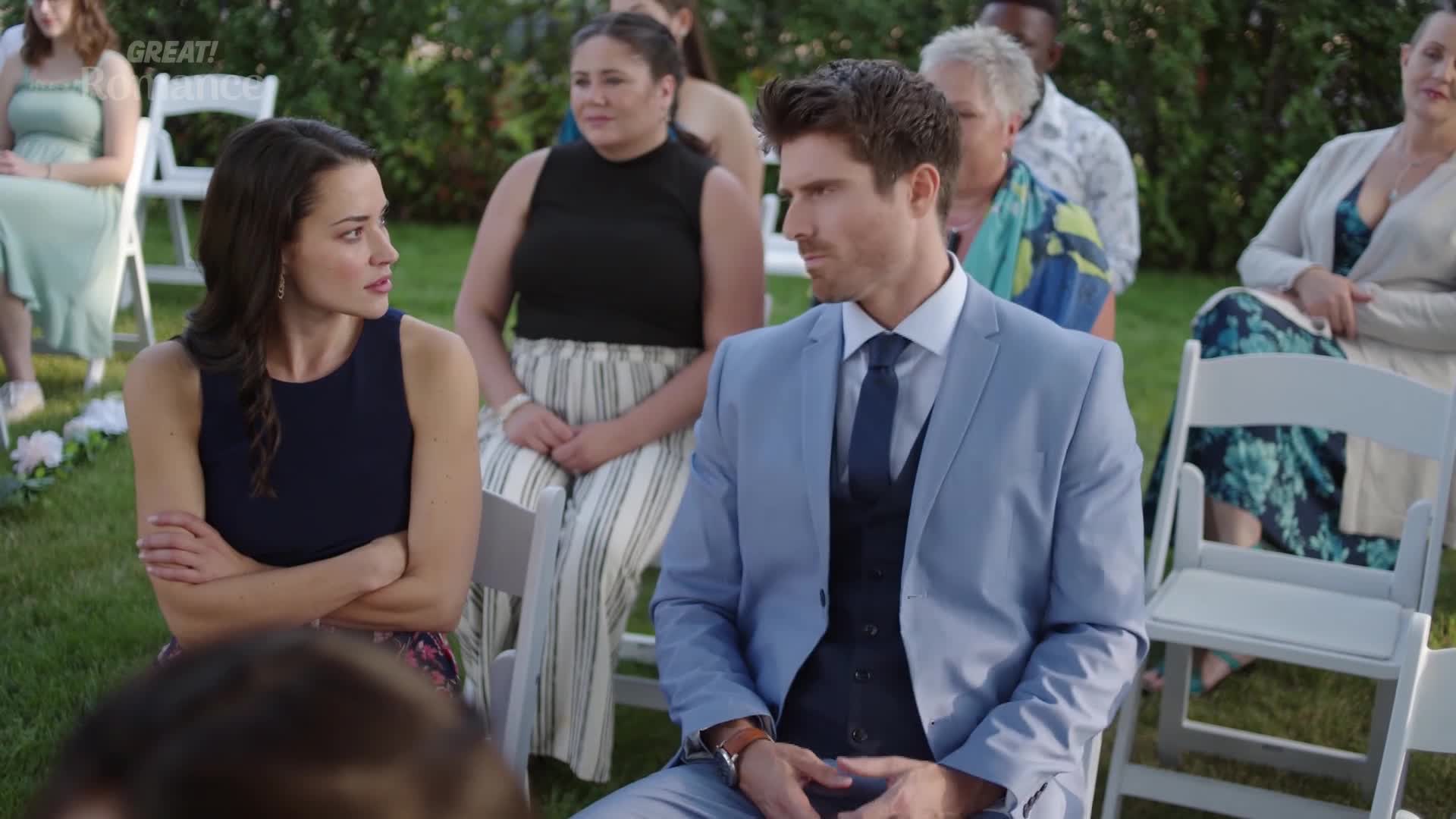 A groom in a light blue suit and a woman in a navy dress sit amongst guests at an outdoor wedding. Their expressions suggest a moment of tension before the ceremony.