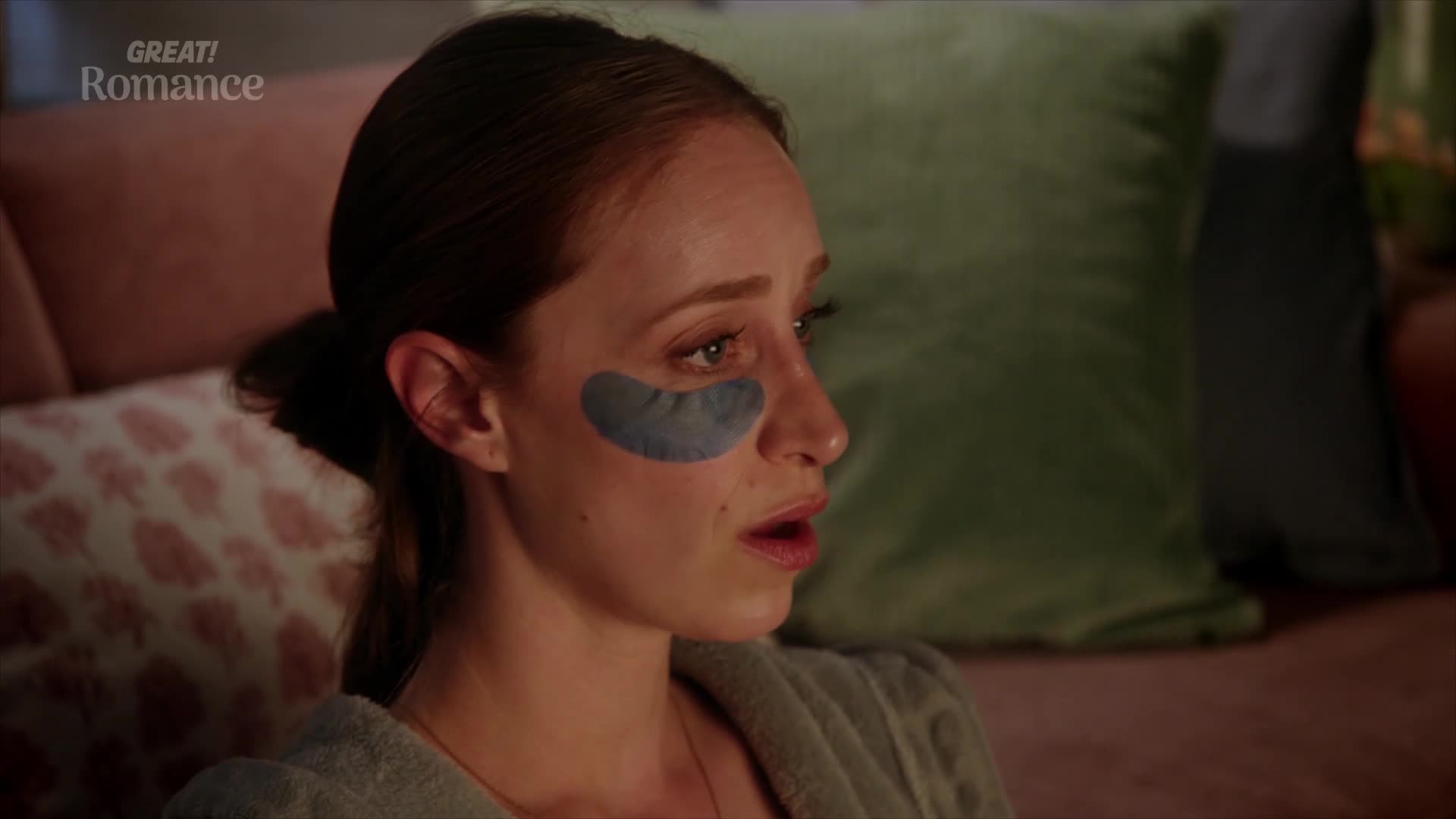 A young woman with grey under-eye patches sits on a sofa, her mouth slightly open. She wears a grey robe, looking off to the side with a concerned expression.