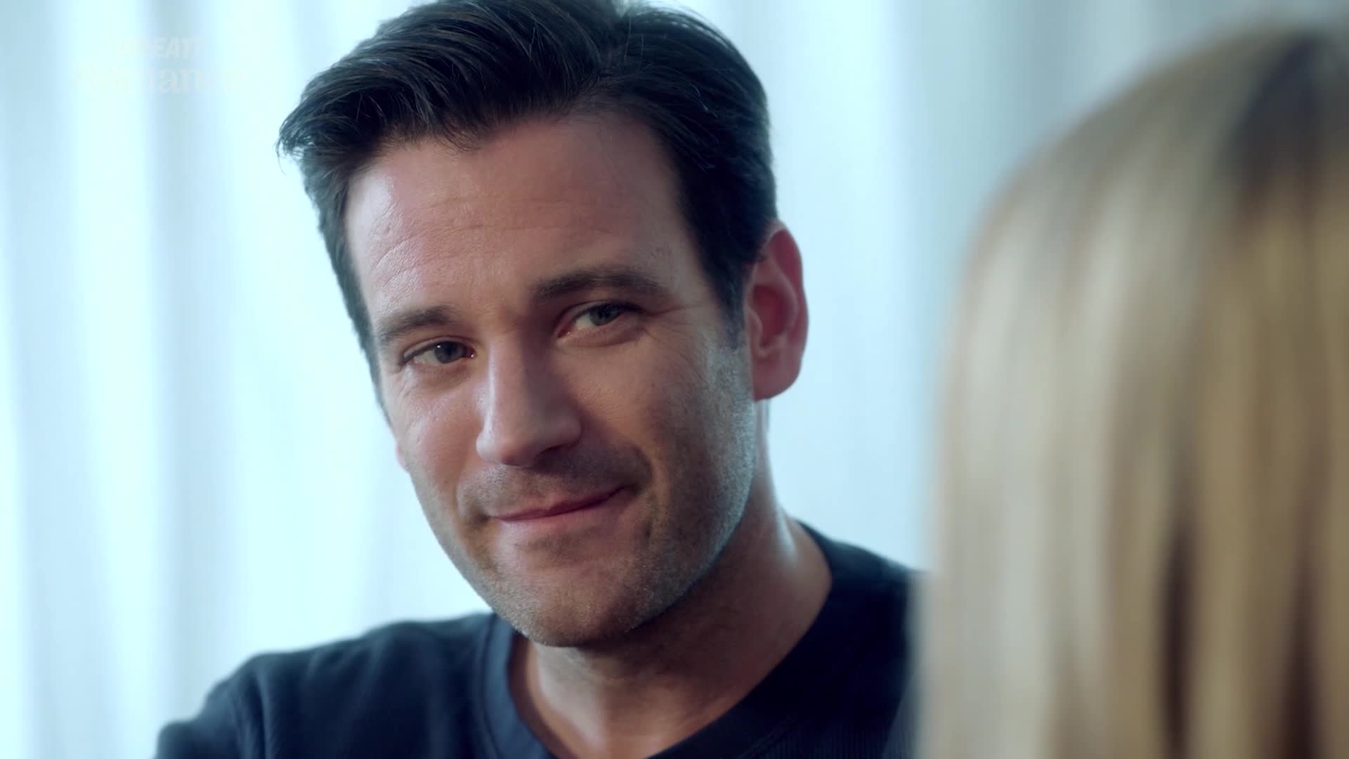 A man's face, framed by dark hair, turns towards the camera with a subtle, knowing smile. His eyes hold a warmth that suggests a shared, intimate moment, perhaps from a quiet scene in a British drama.