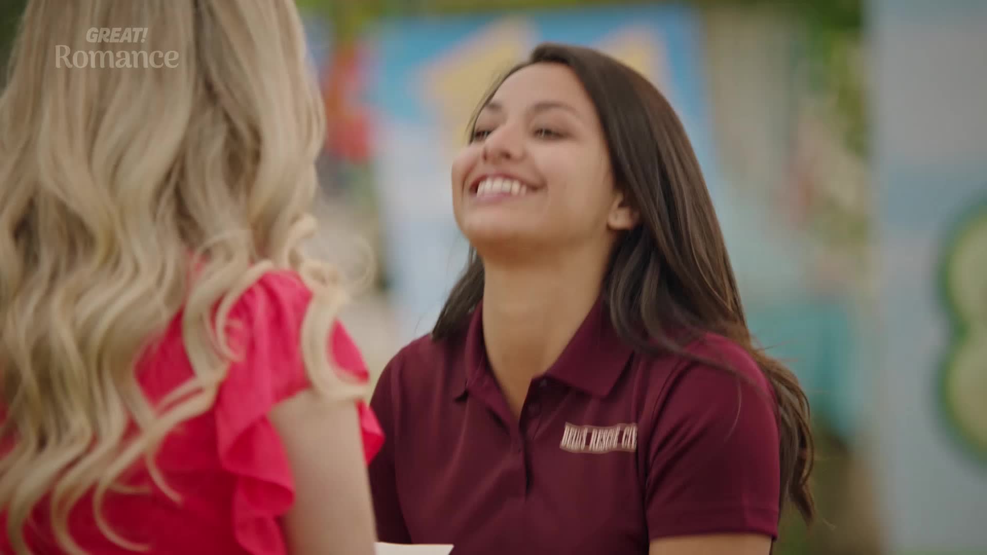 A young woman with dark hair, wearing a maroon polo shirt, beams as she speaks with a blonde woman in a bright red top. The backdrop is a colourful mural, suggesting a pleasant, perhaps even romantic, outdoor setting.