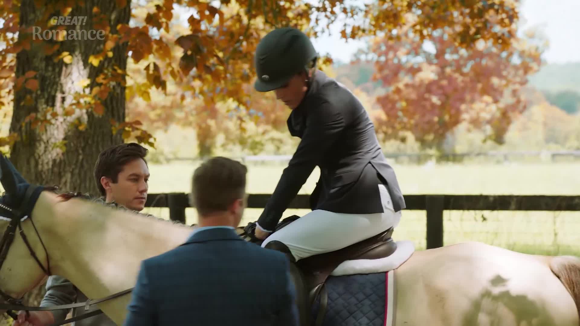 A woman in a riding jacket and helmet is being helped onto a pale horse by two men. The autumn leaves on the trees behind them are a vibrant mix of gold and orange.