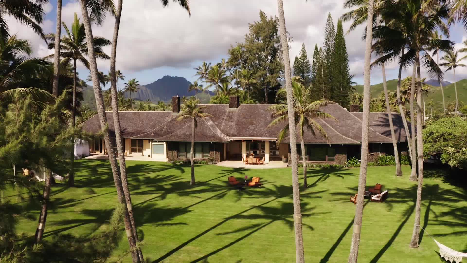 Palm trees cast long shadows across a manicured lawn leading to a sprawling, single-story house. Two orange lounge chairs sit invitingly on the grass, hinting at a peaceful afternoon.