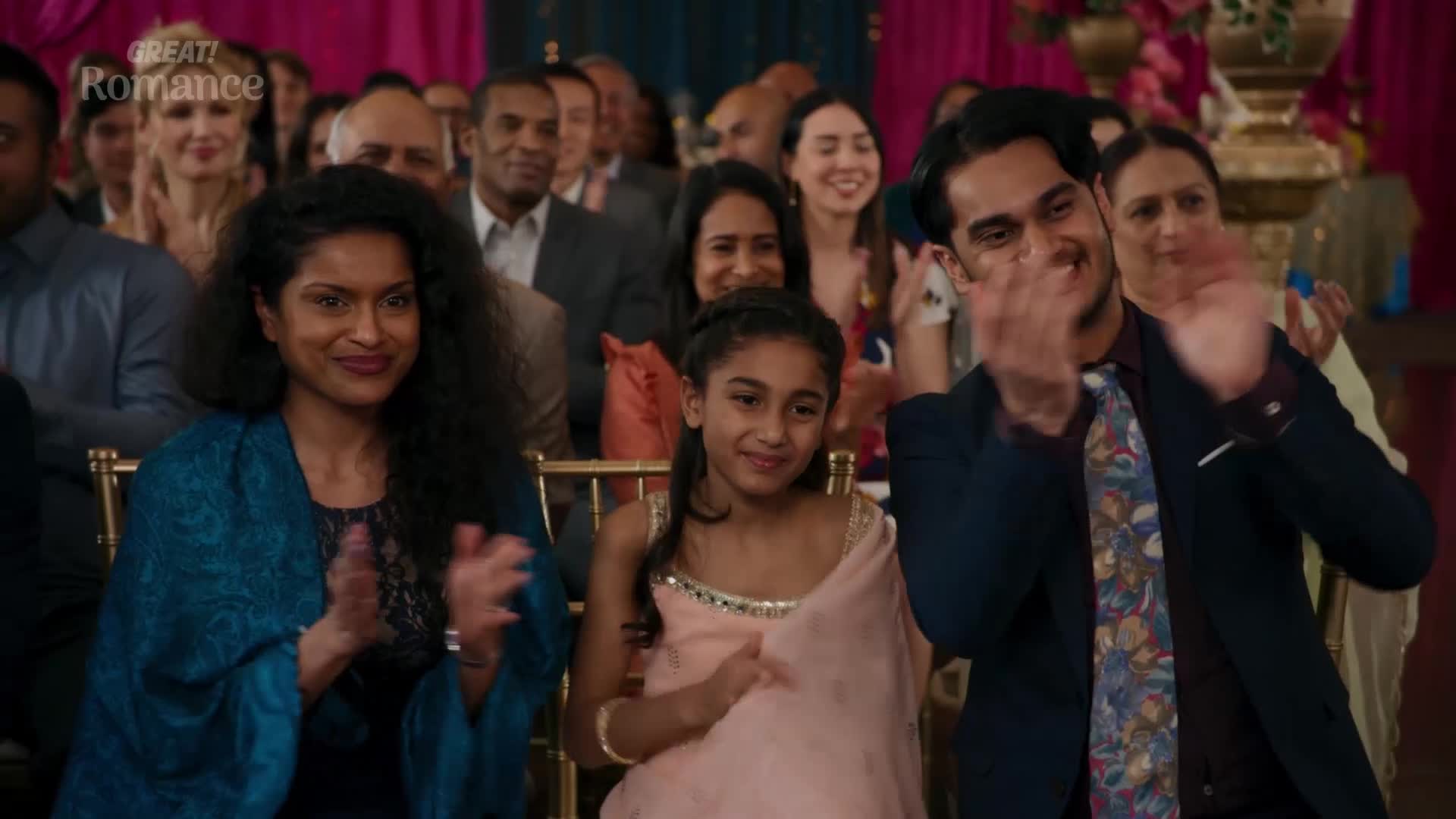 Everyone's clapping, a joyous sound filling the room as a young couple is celebrated. The vibrant colours of the guests' attire suggest a festive occasion, perhaps a wedding reception.