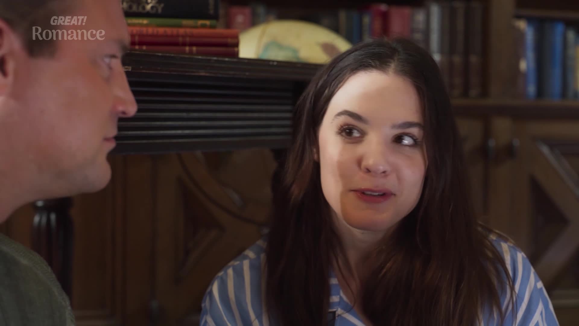 A woman with dark hair, wearing blue and white striped pajamas, speaks animatedly. Beside her, a man in a green shirt listens intently, his profile turned away.