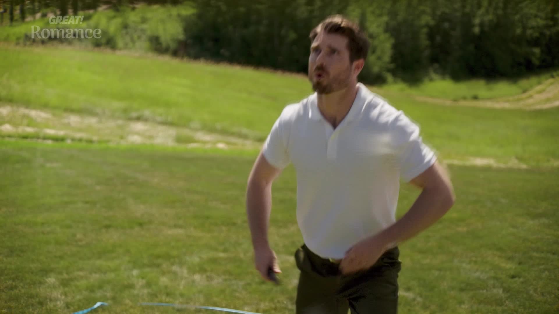 A man in a white polo shirt is looking up, his hands near his waist. A thin blue ribbon stretches across the grass in front of him.