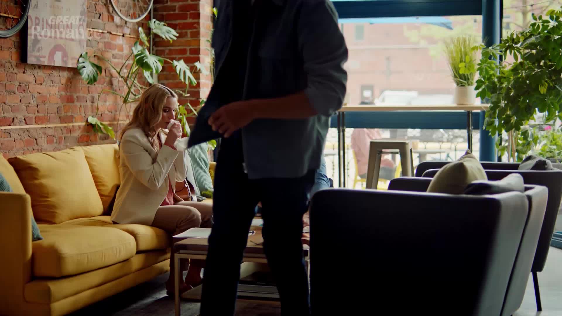 A man in a blue jacket walks past a woman on a yellow sofa. She's holding a tissue to her nose, looking down at some papers.