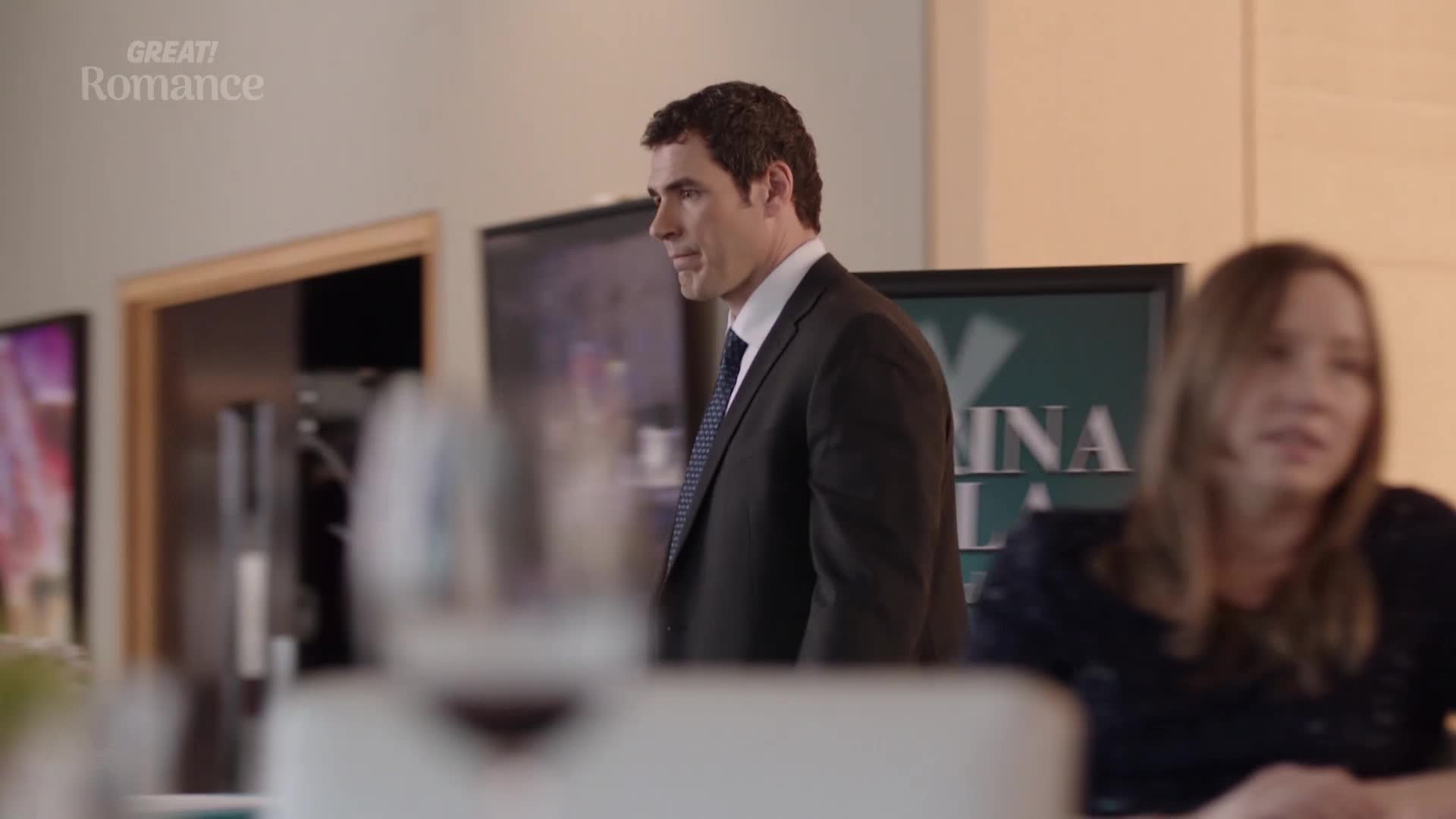 A man in a dark suit and patterned tie walks past a woman seated at a table. The "GREAT! Romance" logo appears in the top left corner.