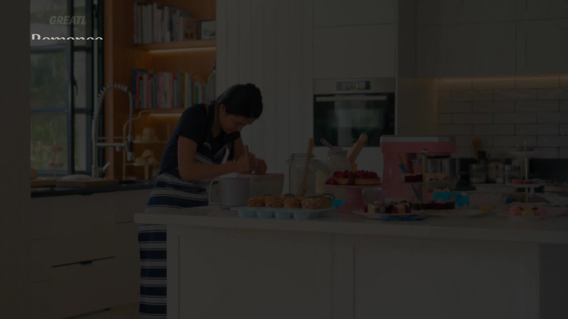 A young woman, wearing a striped apron, is busy whisking something in a bowl on the kitchen counter. A tray of cupcakes sits nearby, ready for decoration.