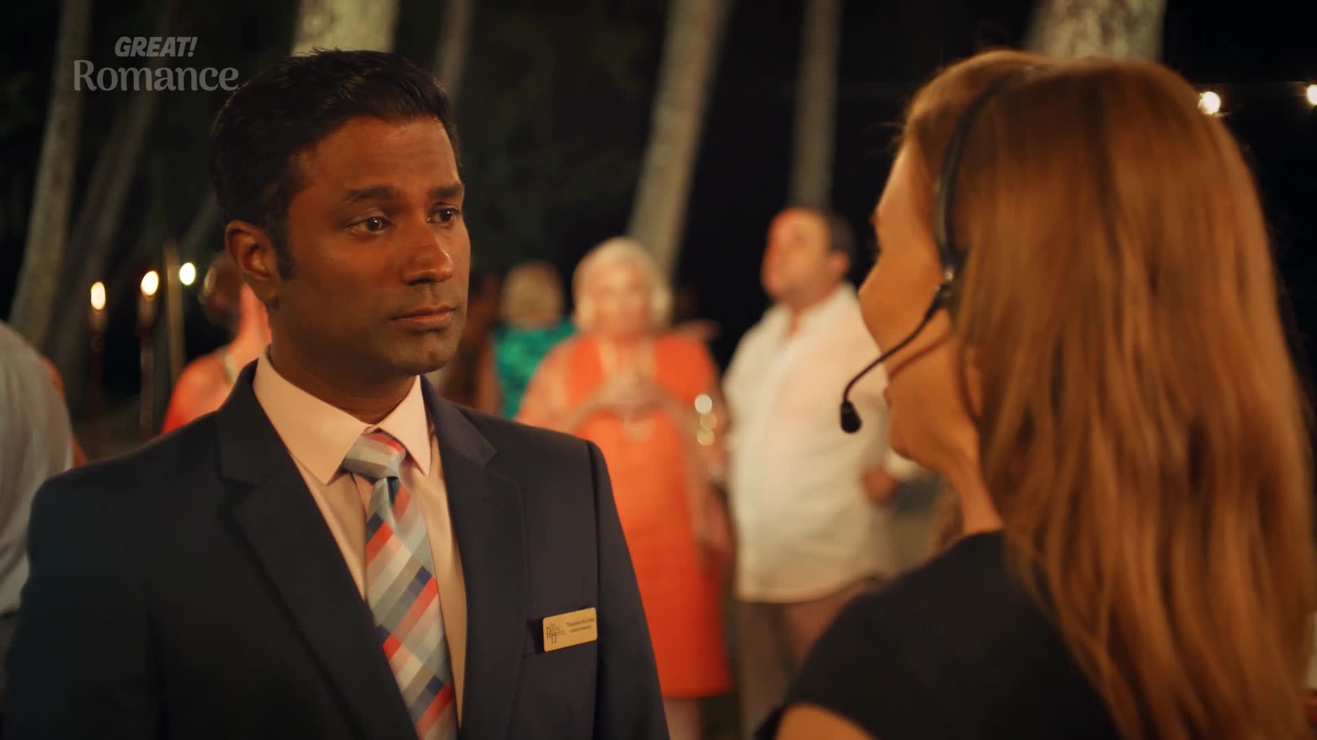 A man in a suit stands attentively, his gaze fixed on a woman with a headset. Behind them, a crowd mingles under soft lighting, suggesting a festive evening event.
