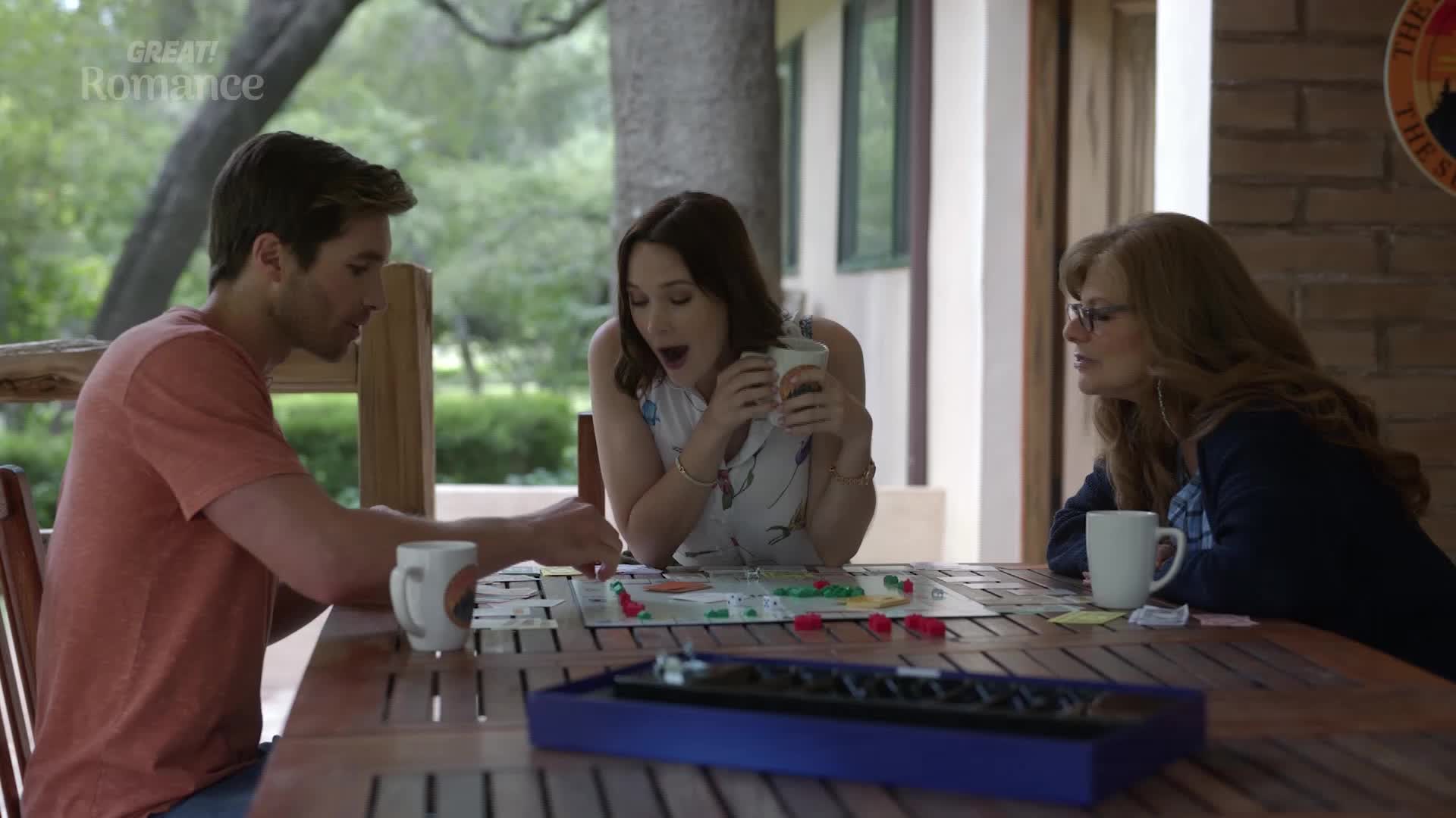A man in a coral t-shirt reaches for a game piece on the board. Beside him, a woman with wide eyes and an open mouth holds a mug, looking surprised by the game's progress. Across from them, a woman with glasses watches the board intently.