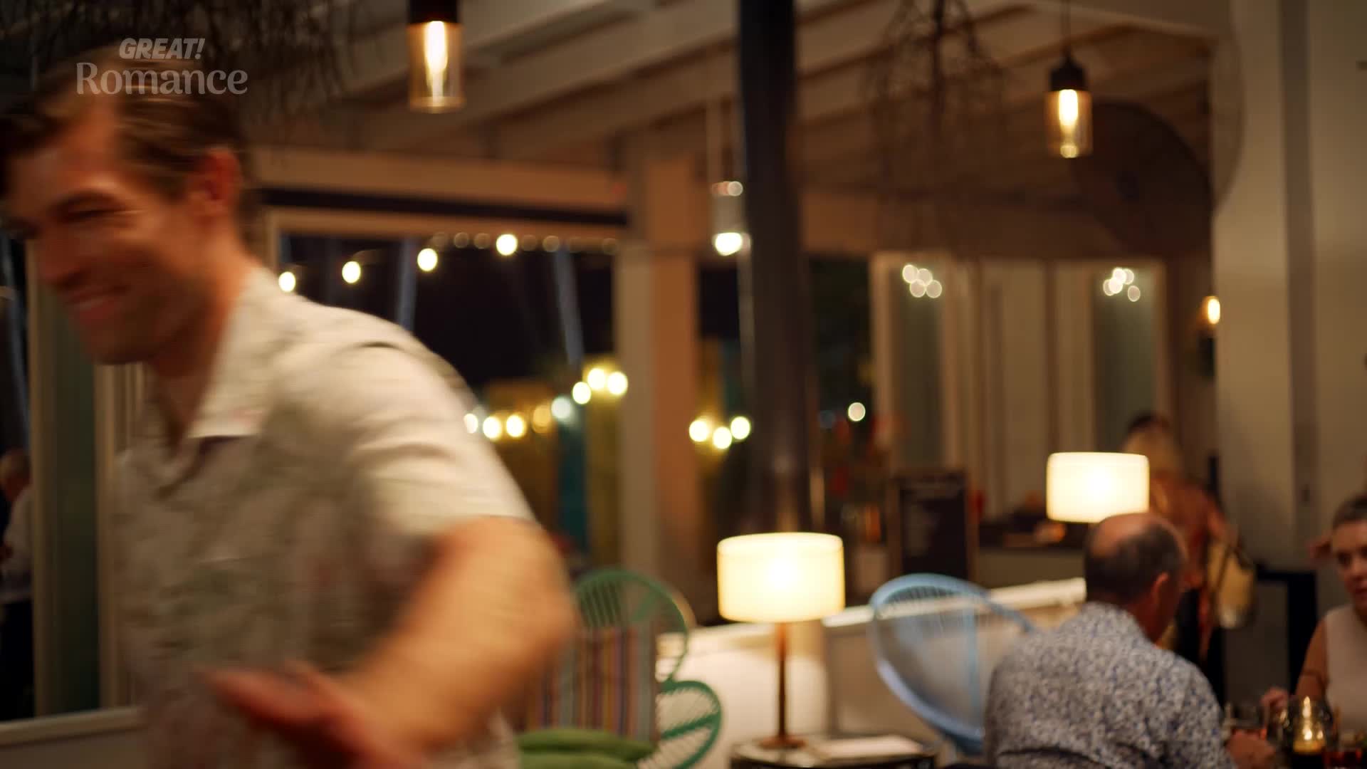 A man in a patterned shirt laughs as he moves through a restaurant. Soft lights glow from lamps and strings of bulbs, illuminating tables where people are dining.