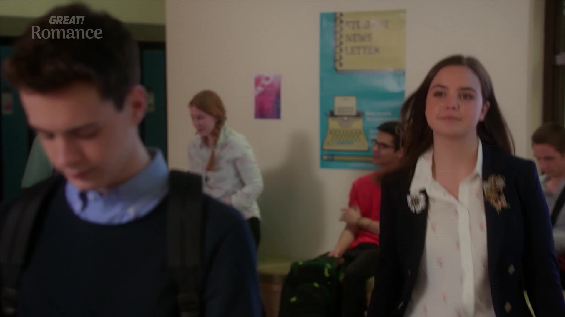 A young woman in a navy blazer walks past, her dark hair falling around her shoulders. Behind her, a boy with a backpack looks down as he moves through the hallway.