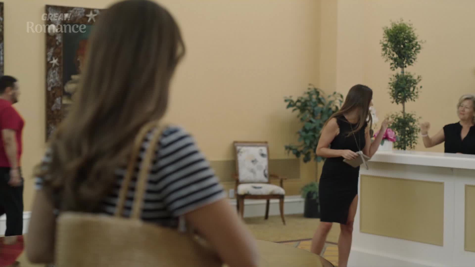 A woman in a black dress approaches the reception desk, her hand outstretched. Another woman, behind the desk, smiles and gestures.